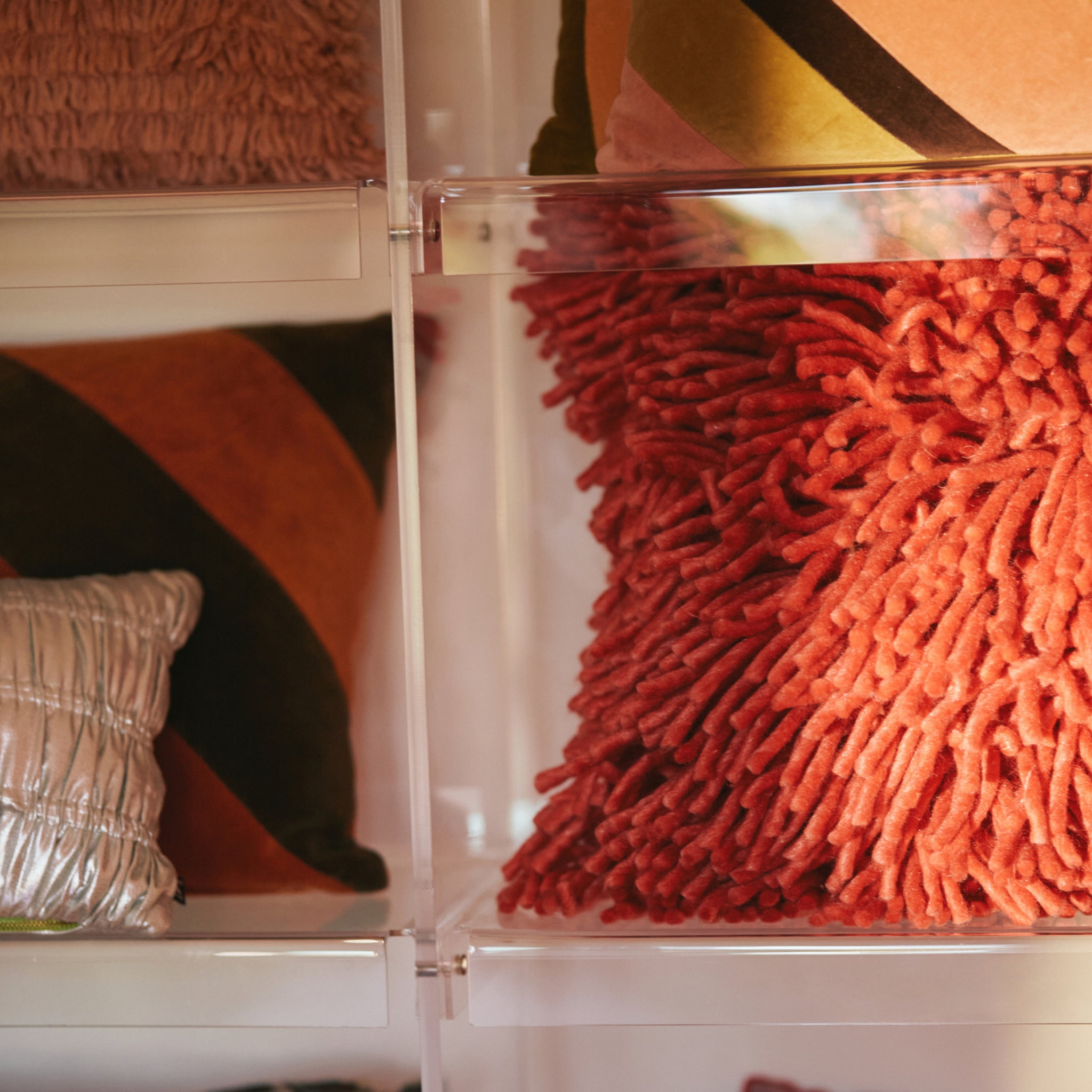 Een close-up van sierkussens op planken, met de HK Living Rugged Kussen Kersenrood 50/50, een glanzend zilver kussen, en HK Living kussens met warme geometrische patronen.