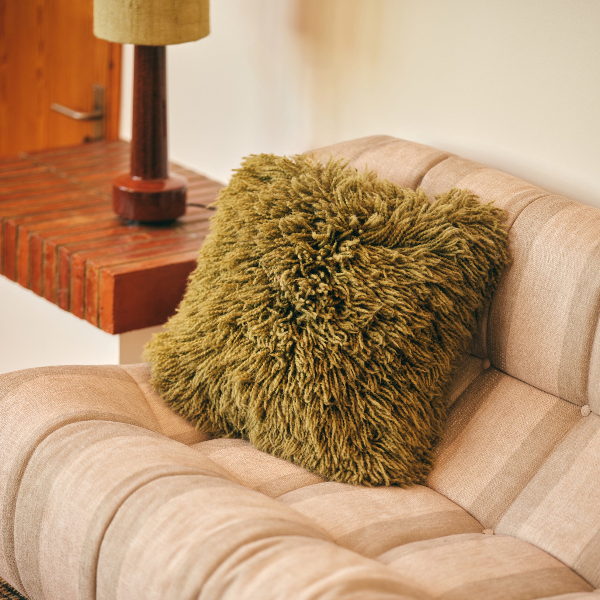 De HK Living Rugged Kussen Olijfgroen 50/50 van HK Living staat op een beige fauteuil naast een houten tafel en lamp en geeft de ruimte een gezellige retro uitstraling.