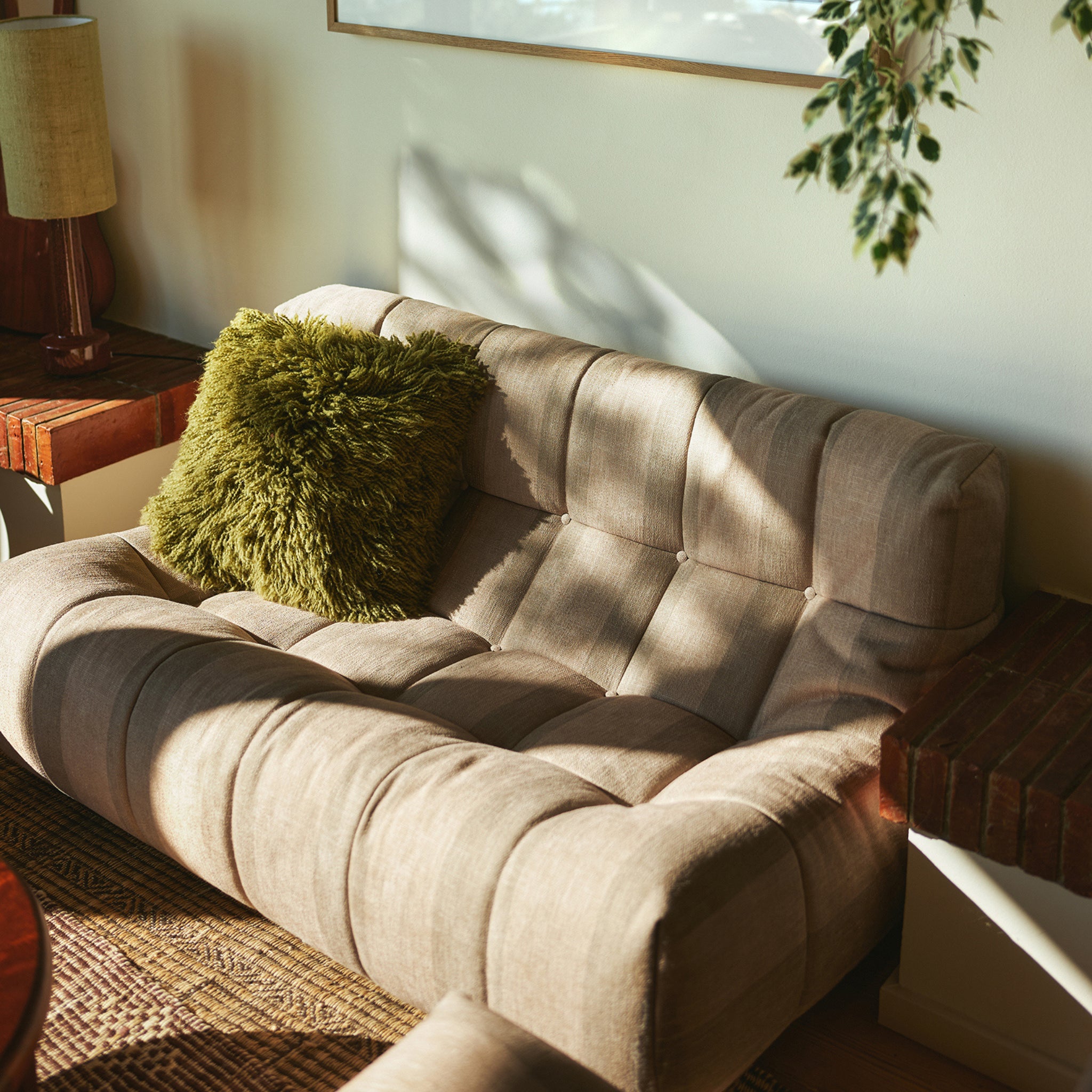 Een beige getufte loveseat met een HK Living Rugged Kussen Olijfgroen 50/50 kussen staat in een zonovergoten hoek, naast houten bijzettafeltjes en een hangende bladplant.