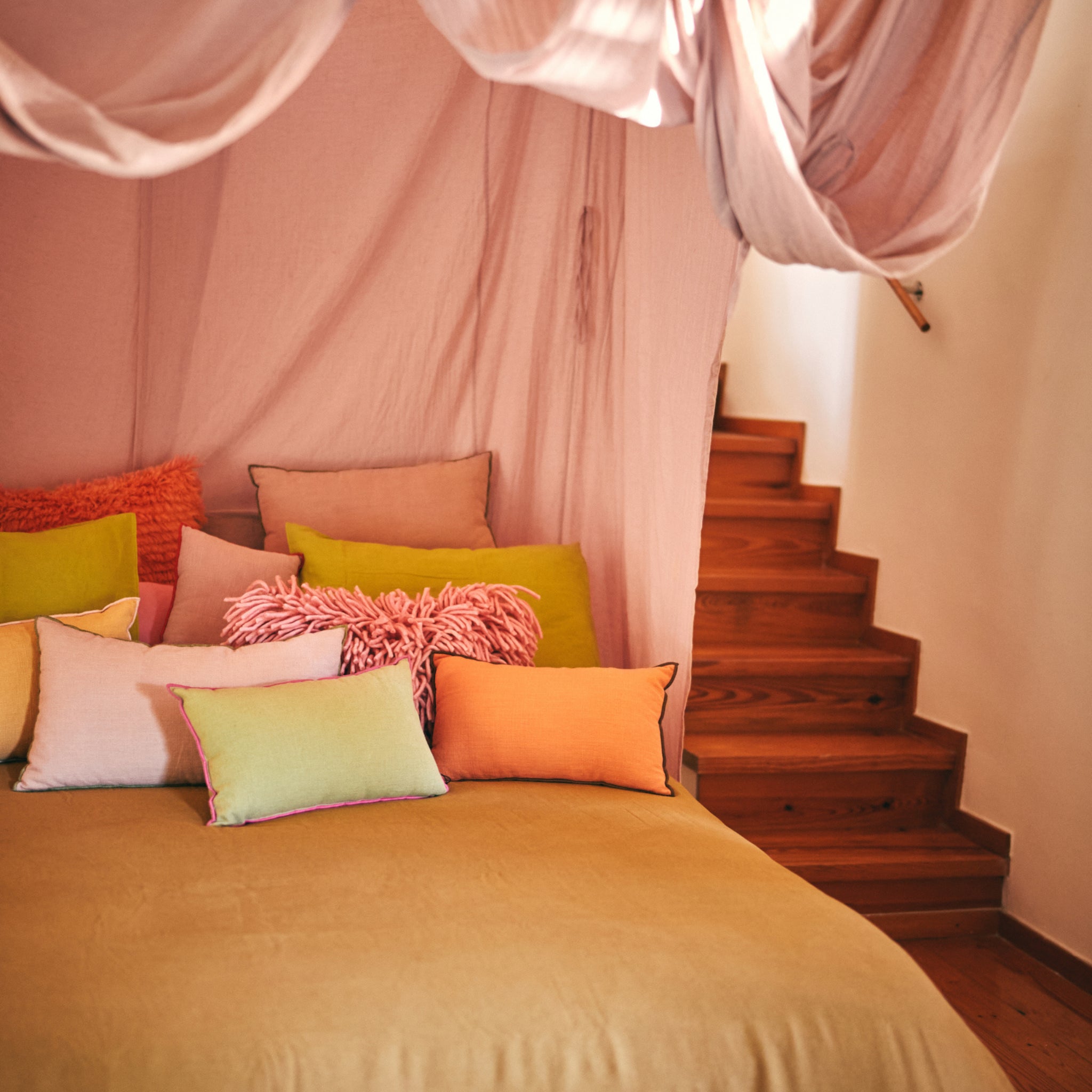 Een knus bed met een beige hoes, kleurrijke kussens waaronder de HK Living Kussen Mansion Lichtgroen 50/30, en een roze hemel erboven staat bij een houten trap. Warme verlichting van HK Living creëert een zachte, uitnodigende sfeer.