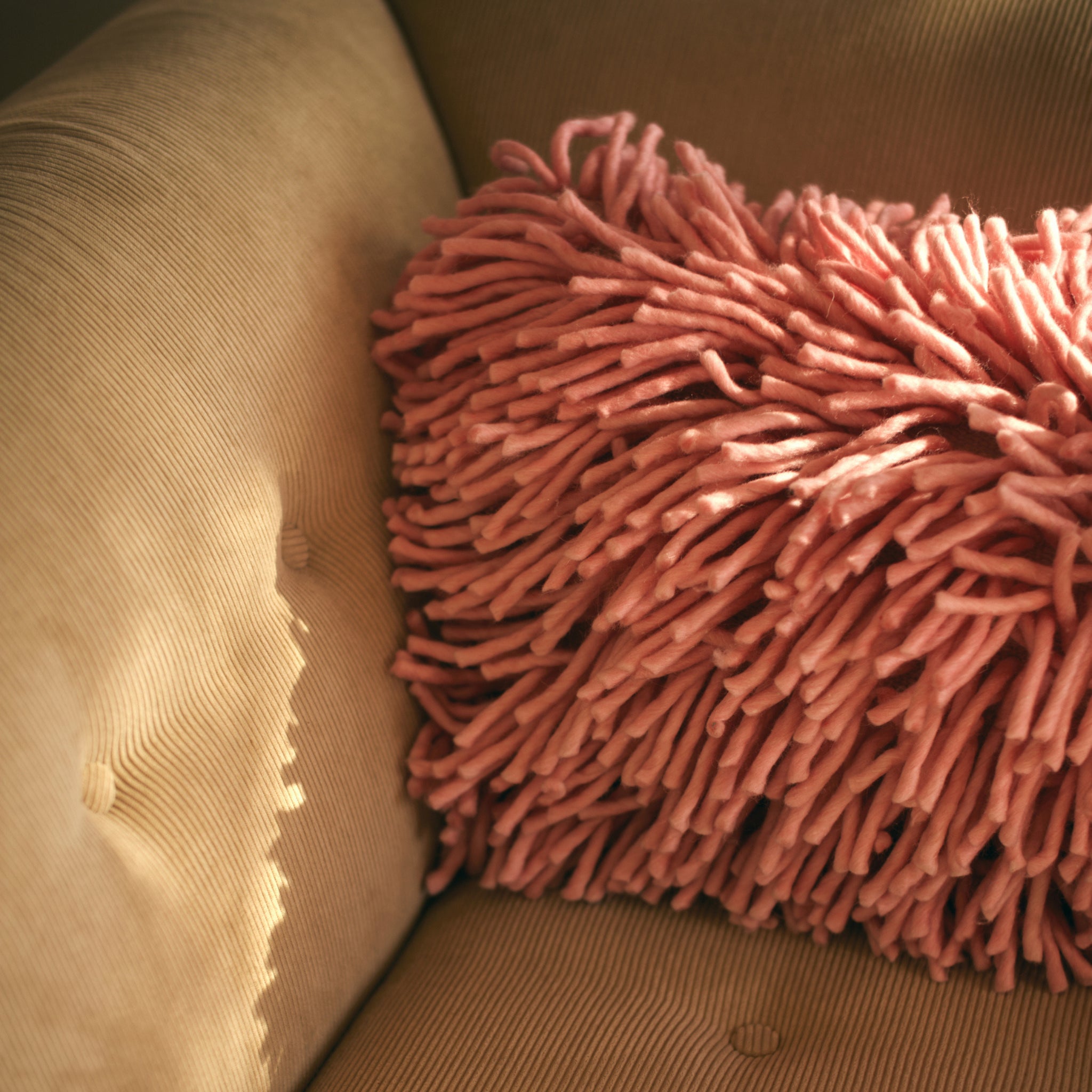 Het HK Living Rugged Kussen Roze 70/45 kussen voegt een vleugje roze toe aan een bruine corduroy fauteuil, terwijl zacht zonlicht zachte schaduwen creëert op het oppervlak van de stoel.