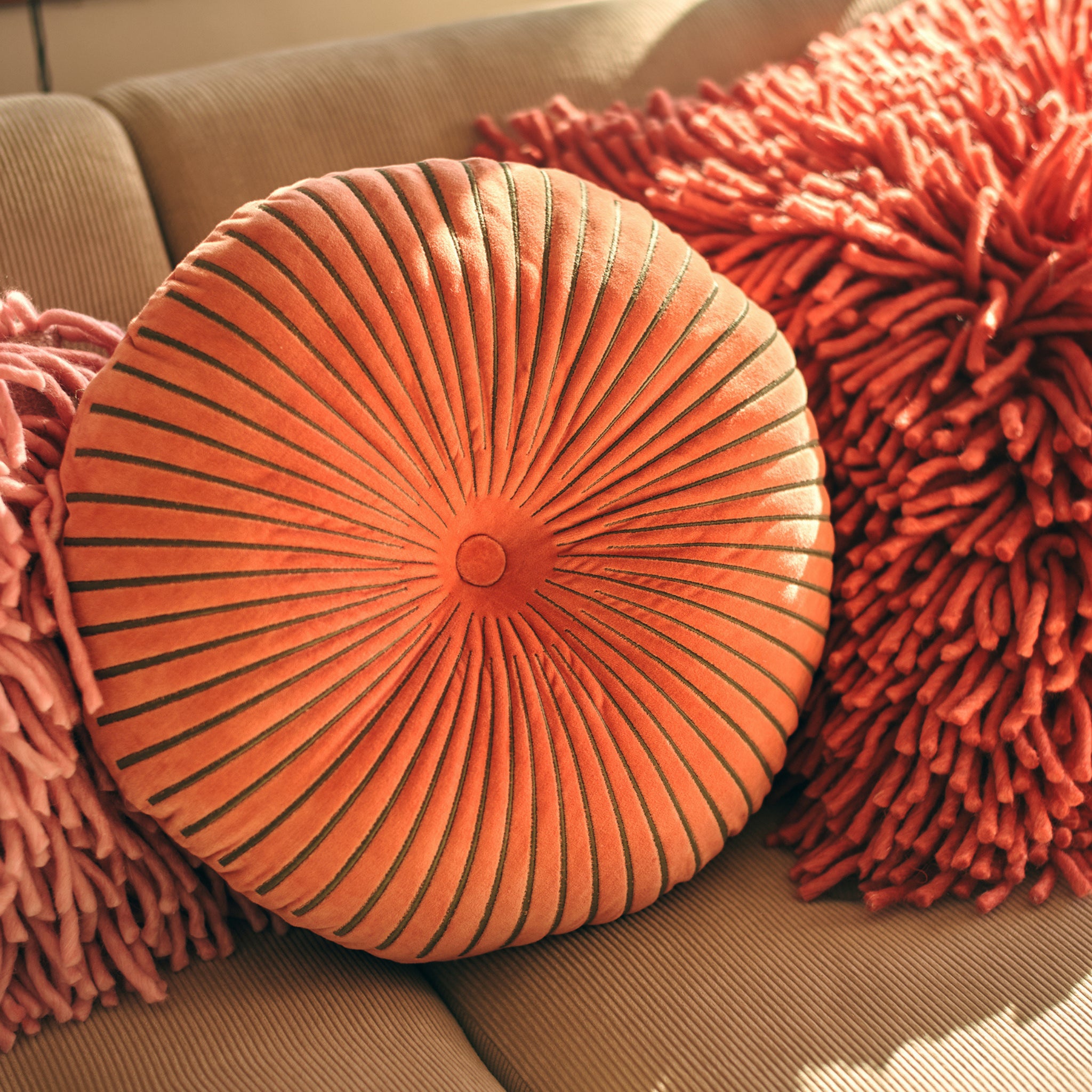 De HK Living Ripple Rond Kussen Sienna D50, een rond kussen van oranje fluweel met stralende plooien, rust op een beige bank tussen twee roze shaggy kussens, alles badend in warm zonlicht voor een gezellige ambiance.
