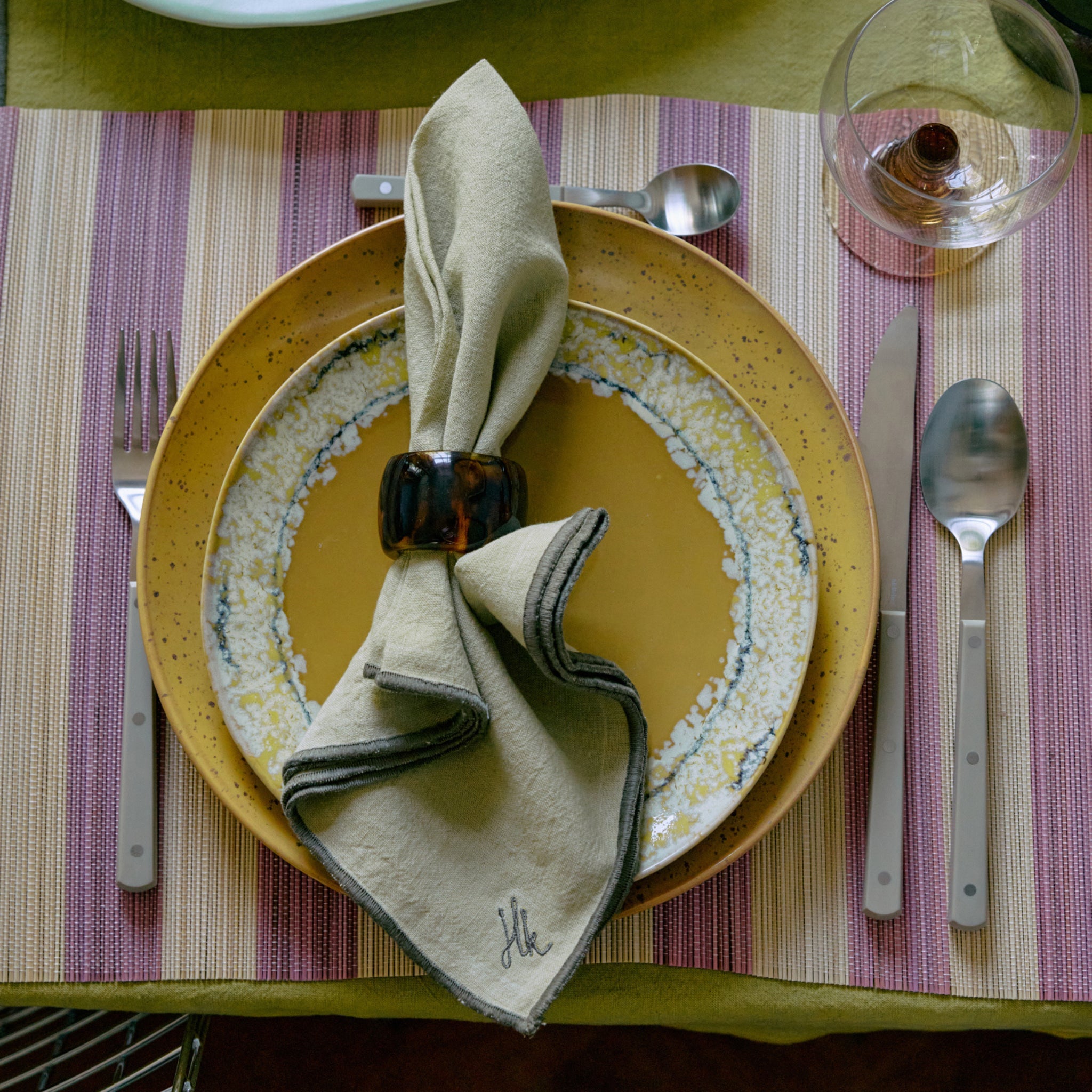 Een stijlvolle tafel is voorzien van een geel bord, een gedessineerd bijzetbord, een gevouwen servet met de HK Living Servetring Sunrise 4 st., zilverwerk op een gestreepte placemat en een helder glas voor een eigentijdse tafeldekking.