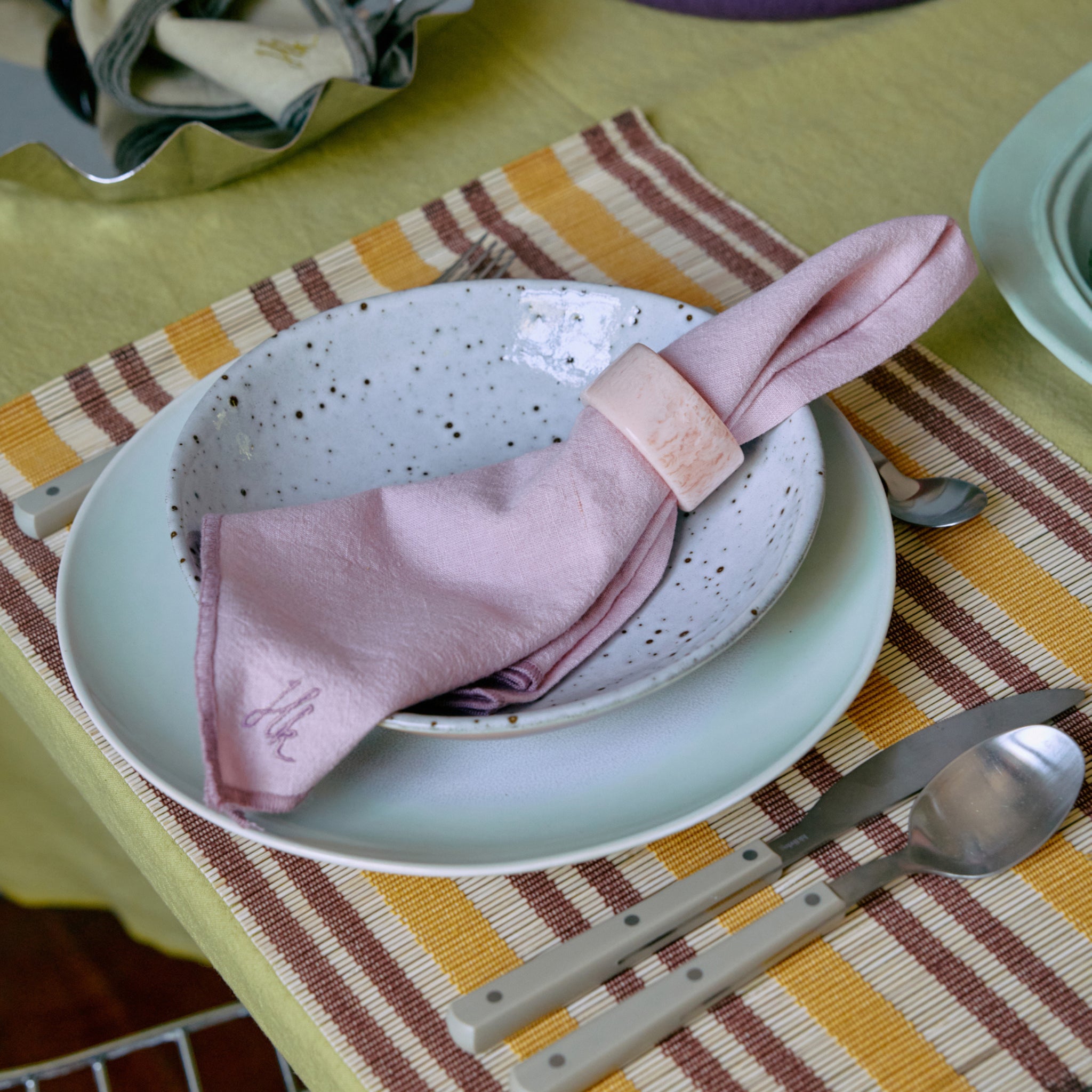 Een couvert met een gespikkelde kom op een lichtgroen bord, een roze servet in een HK Living Servetring Sunrise en bestek op een geelbruin gestreepte placemat - perfect voor een eigentijdse tafeldekking.
