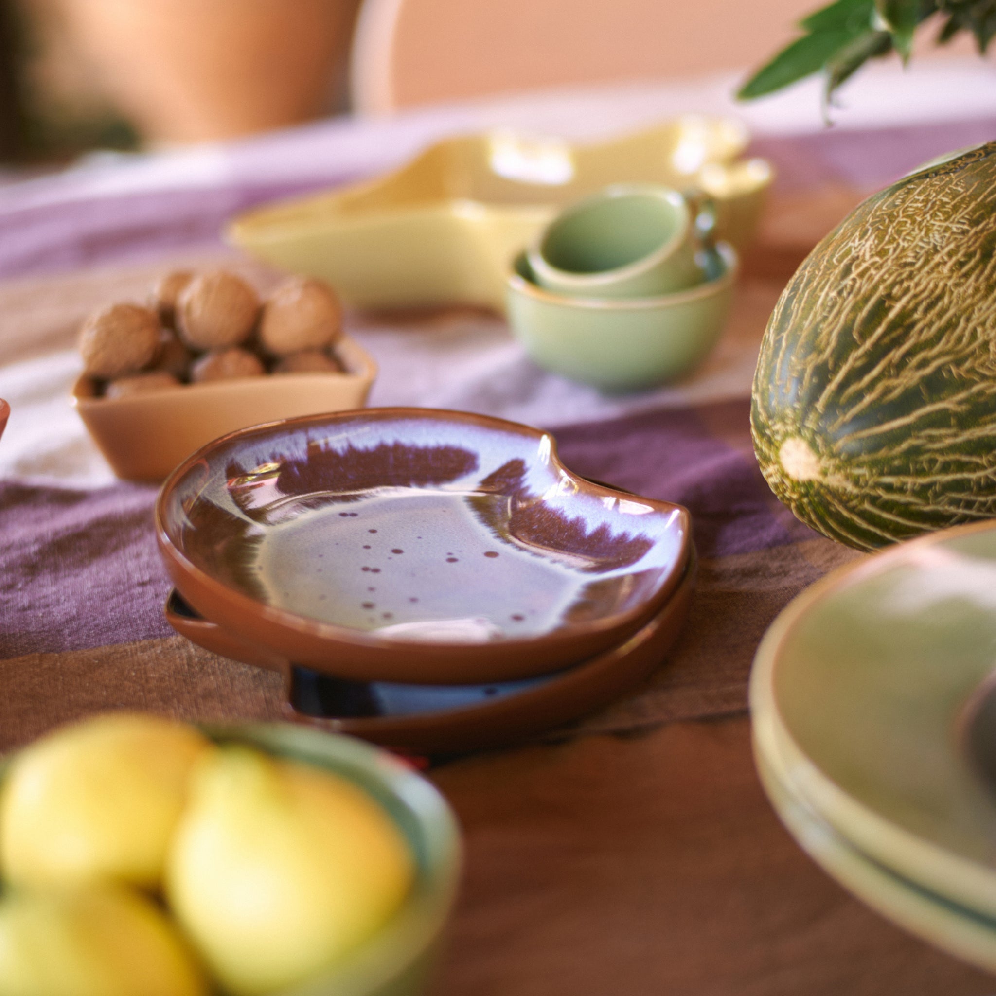Keramische schalen, een HK Living Shell Schaal Sienna en schaaltjes met noten omringen een meloen op een gestreept tafelkleed. Citroenen in een schaal en groene bladertakken brengen kleur in de ontspannen, natuurlijke omgeving.