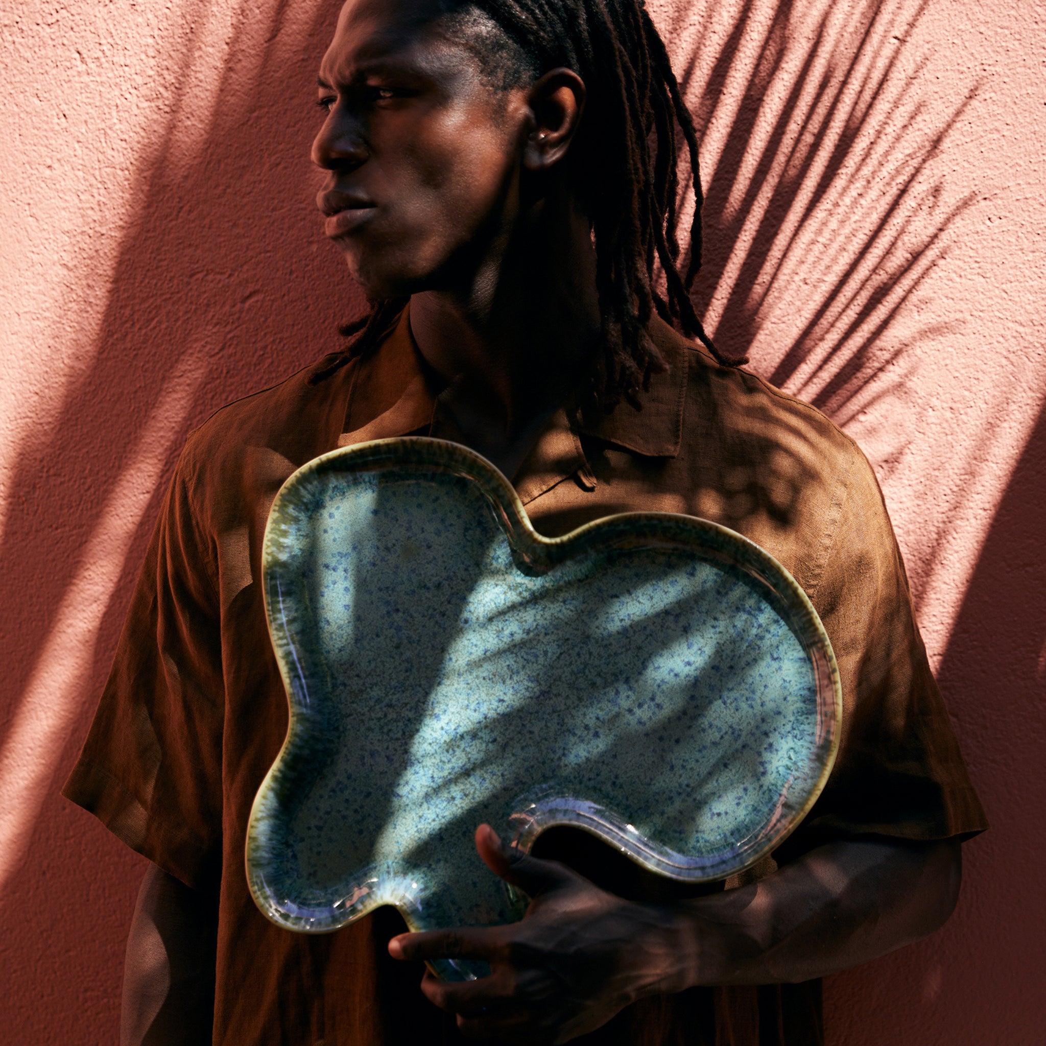 Een man in een bruin overhemd staat tegen een roze muur en houdt de HK Living Shell Schaal Teal vast. Schaduwen van palmbladeren worden over hem en de muur geworpen, waardoor een dynamisch patroon ontstaat.