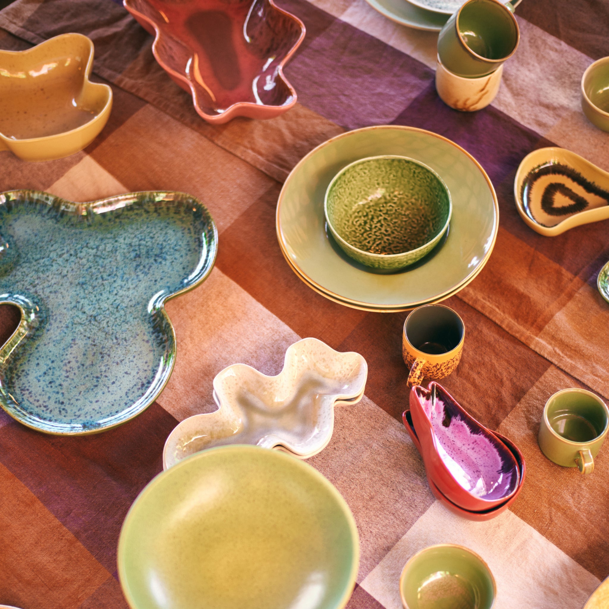 Kleurrijke keramische borden, kommen en kopjes, waaronder een blauwgroen servies en de HK Living Shell Schaal Teal, zijn gerangschikt op een geruit bruin en paars tafelkleed, waardoor een levendige en artistieke tafelsetting ontstaat.