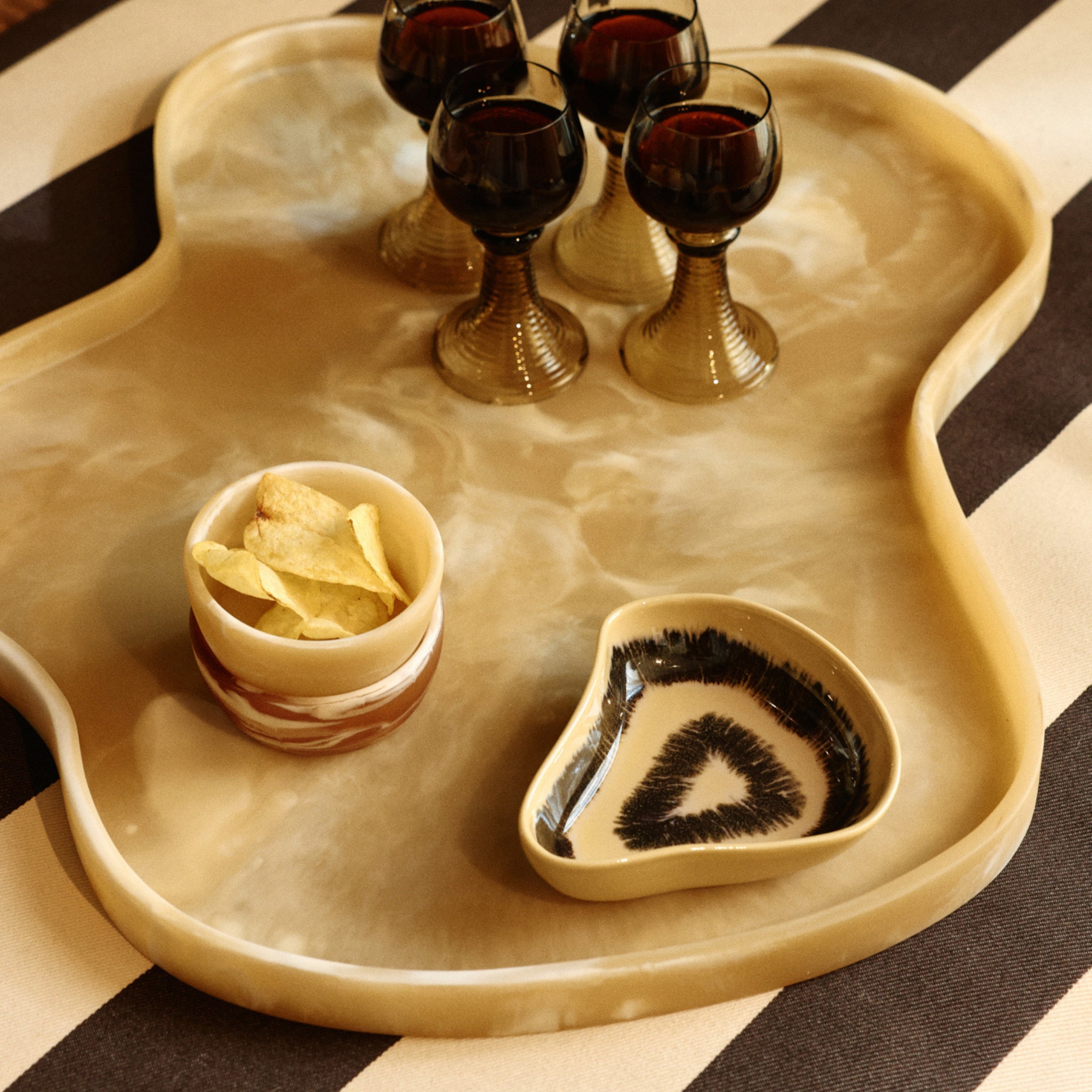 Een beige gemarmerd dienblad bevat vijf glazen donkere vloeistof, een HK Living Shell Tapas Kom Triffle en een schaal met een zwart-wit abstract keramiekpatroon, allemaal op een gestreept oppervlak.