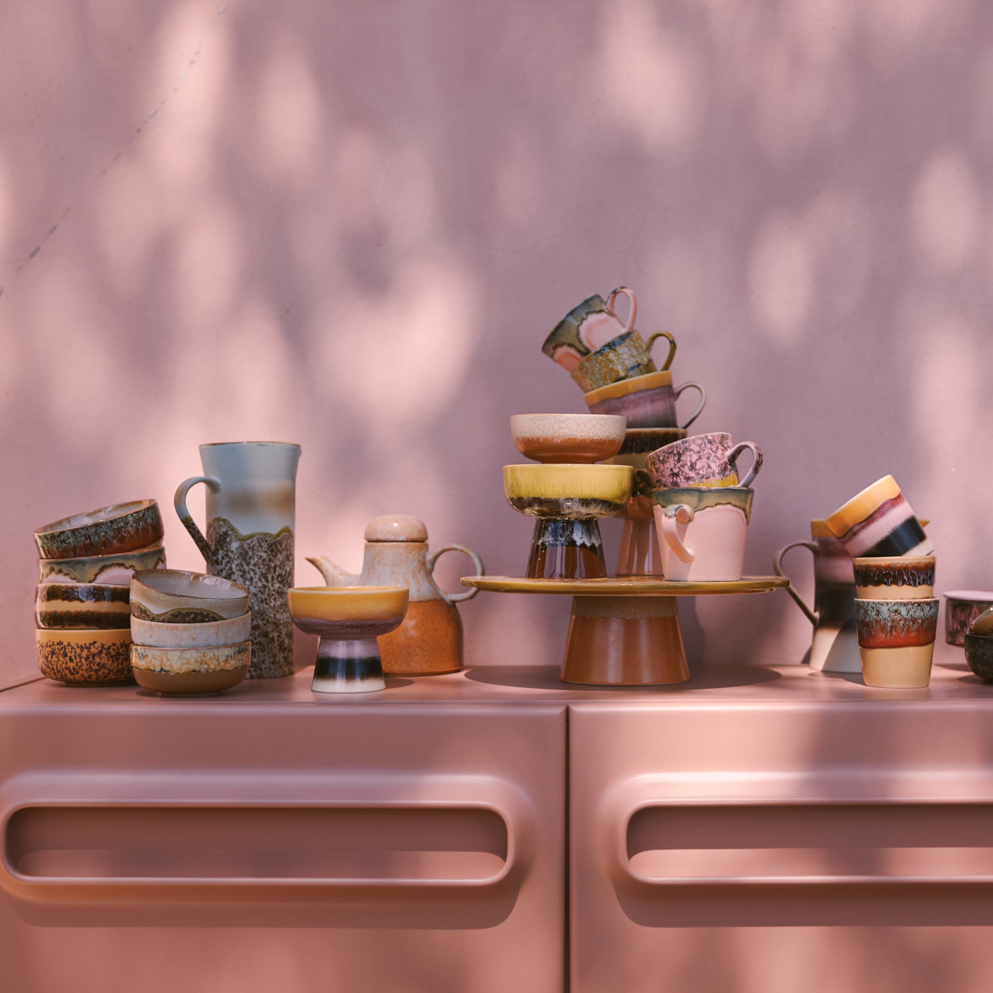 Een stapel HK Living 70s Ceramics Koffiekop Cove mokken geeft een kleurrijk retro tintje aan een roze dressoir tegen een zonovergoten roze muur.