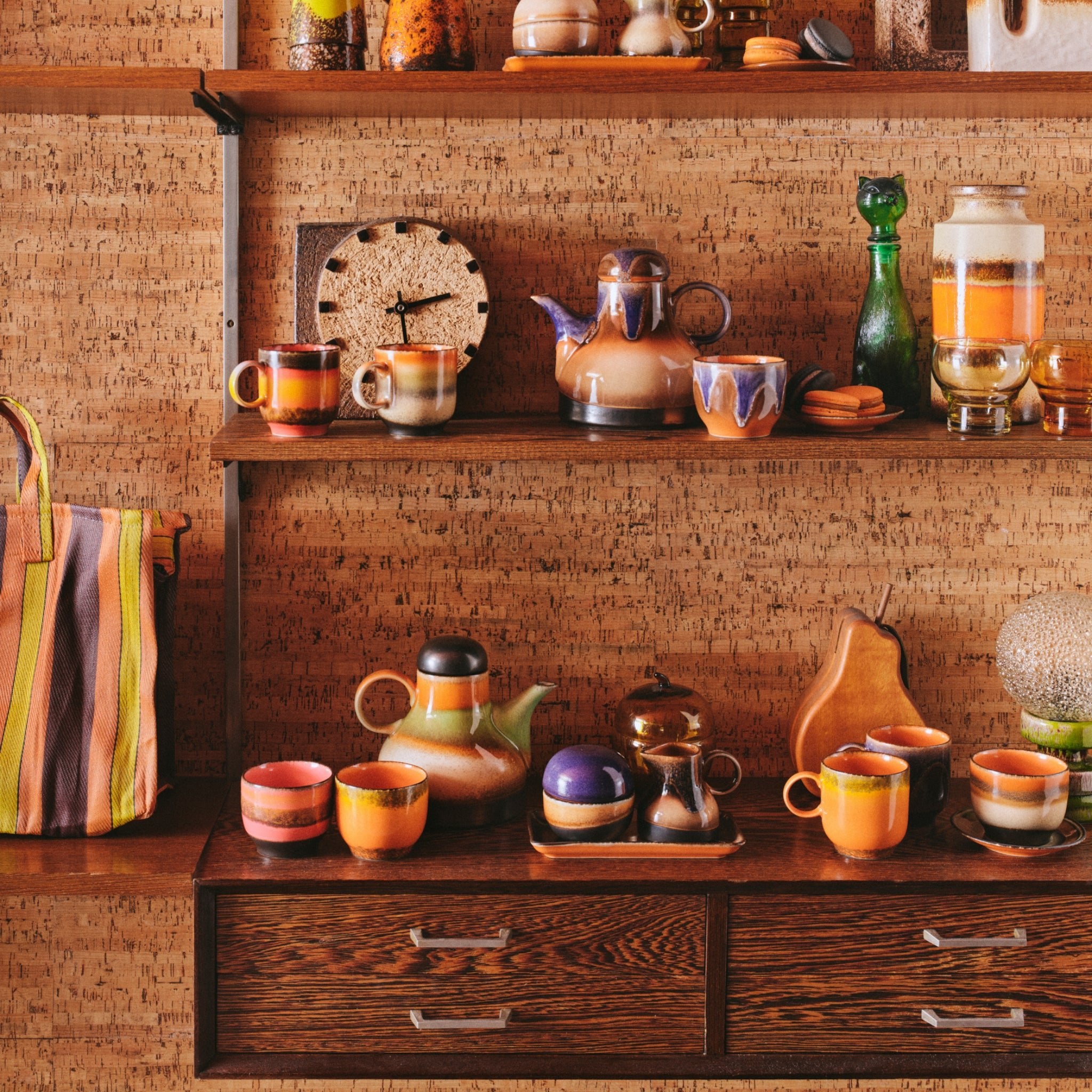 Op een houten plank staan HK Living 70s Ceramics mokken, het HK Living 70s Ceramics Melkkannetje 4PM, theepotten en kommen in aardse en oranje tinten met een gestreepte tas, klok en decor tegen een structuurkleurige muur.