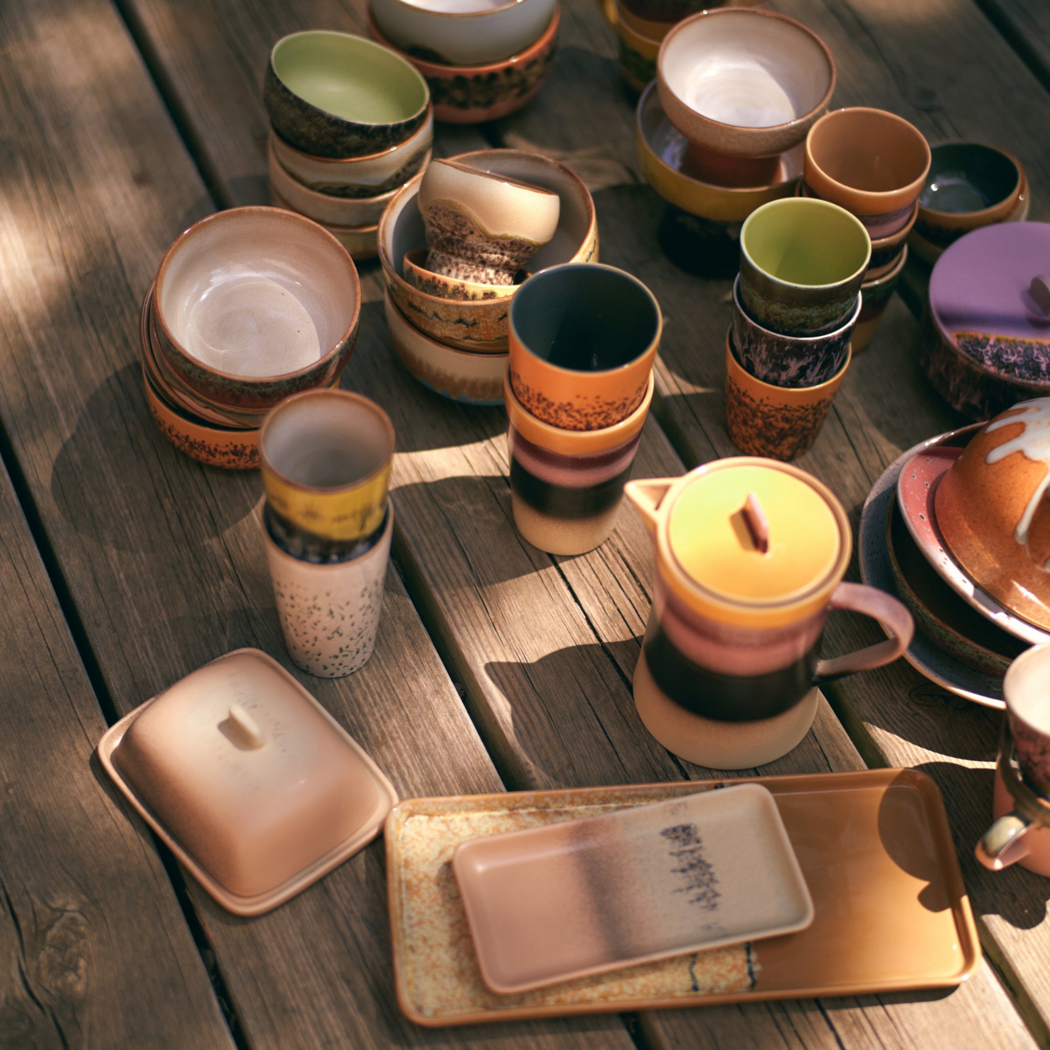 Allerlei keramische mokken, kommen en borden in aardetinten - plus het HK Living 70s Ceramics Botervlootje Breeze - staan buiten op een houten tafel, met zonlicht en schaduwen die een warme, uitnodigende sfeer creëren.