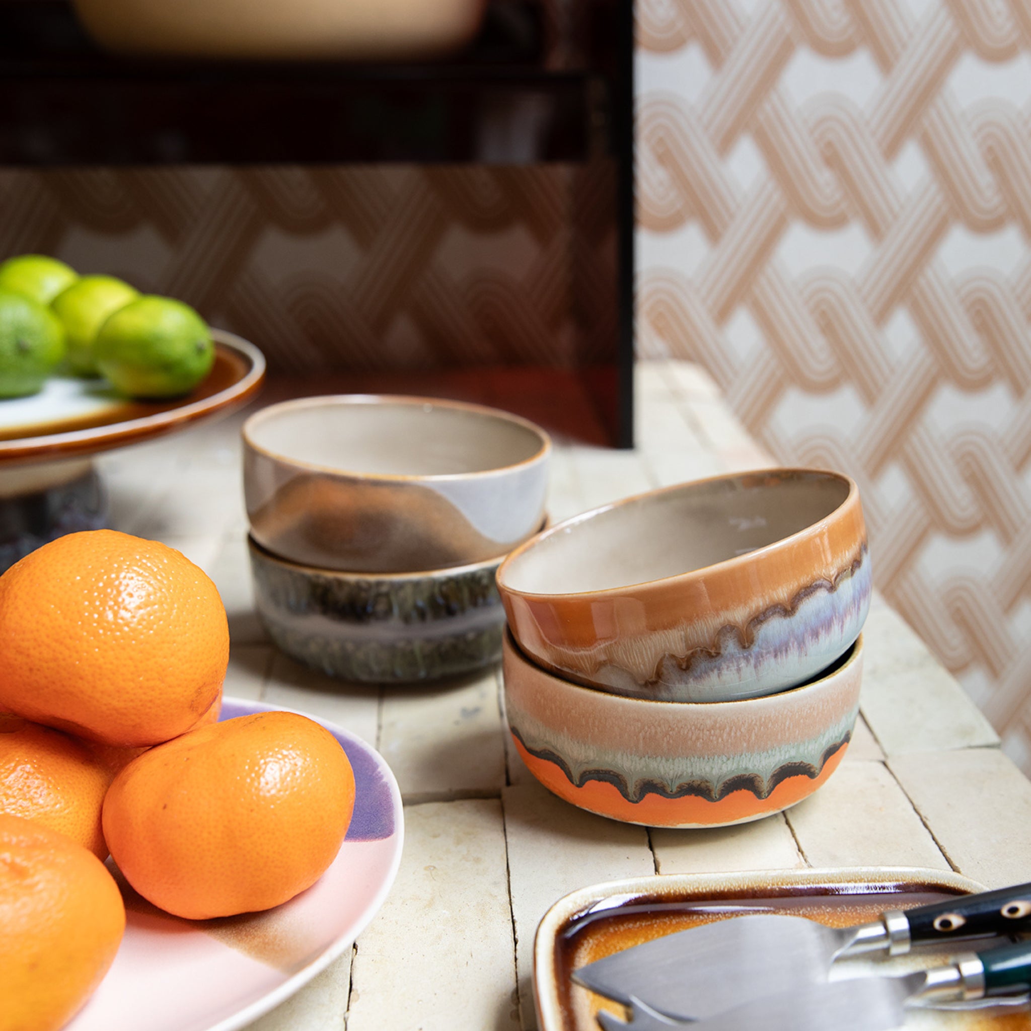 Drie gestapelde HK Living 70s Ceramics Dessertkom Reef kommen, aards met golvende patronen, zijn gecombineerd met heldere sinaasappels en limoenen, tegen een geometrisch behang - een voorbeeld van de kenmerkende stijl van HK Living.
