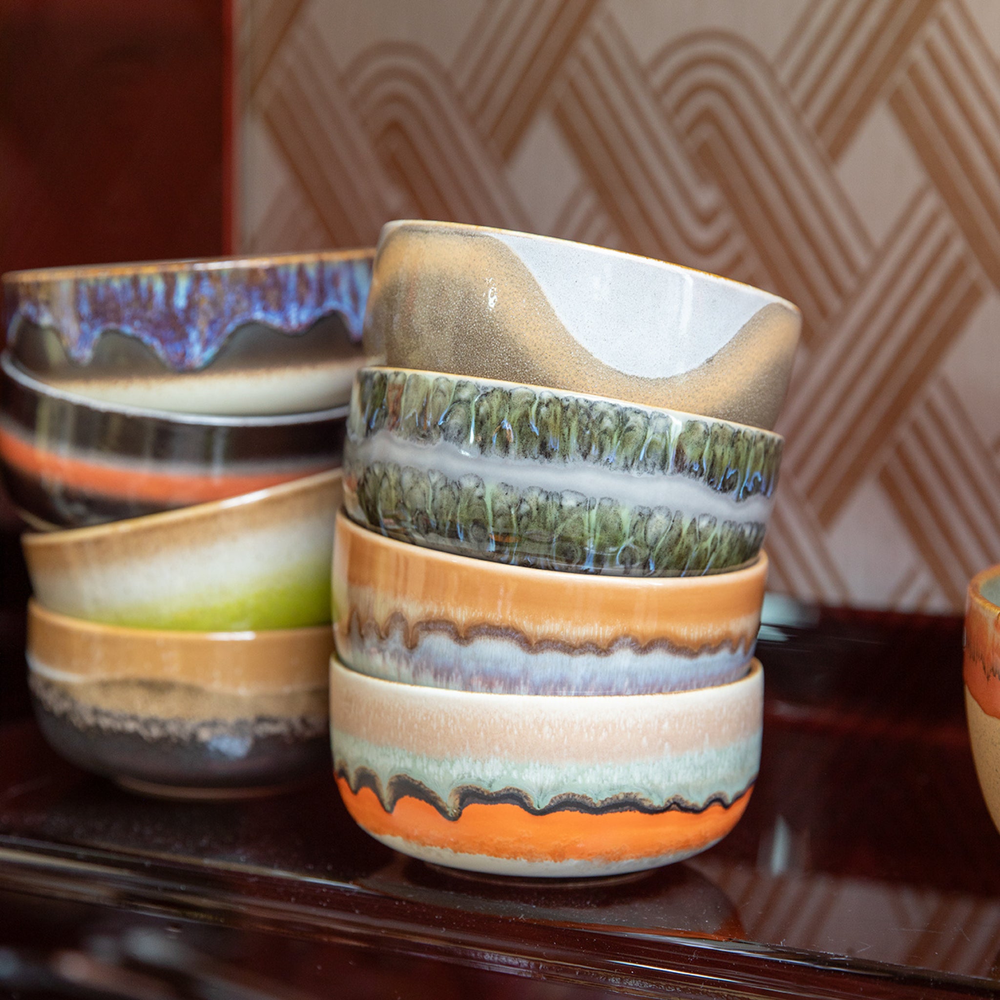 Een stapel levendige HK Living 70s Ceramics Dessertkom Reef kommen (set van 4) heeft unieke glazuurpatronen en staat op een donker oppervlak met een achtergrond met geometrische patronen.