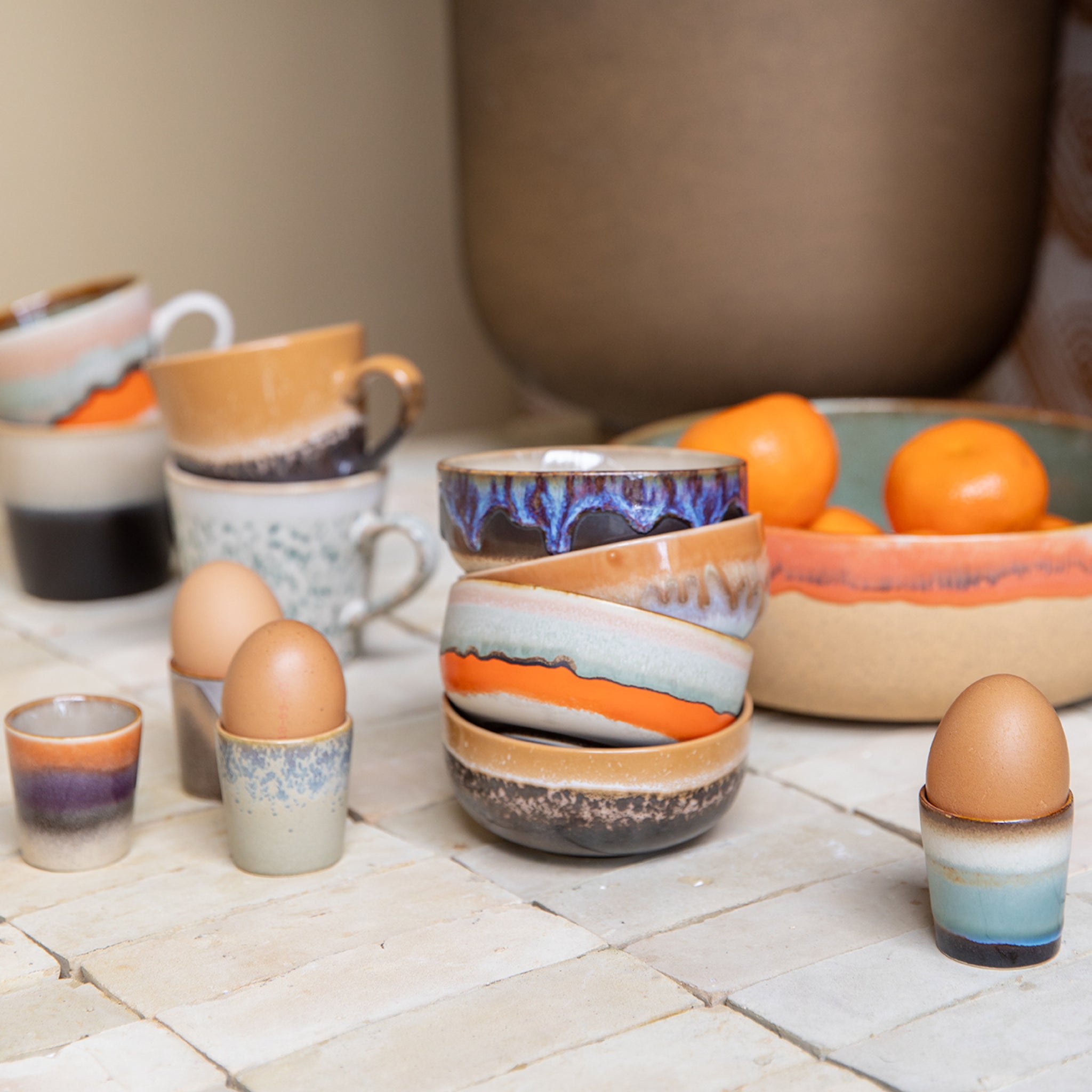 Een levendig display van kleurrijke keramische kopjes, kommen en HK Living 70s Ceramics Eierdop Granite (4 stuks) houdt bruine eieren vast op een betegeld oppervlak. Een schaal met heldere sinaasappels op de achtergrond maakt het levendige arrangement compleet.