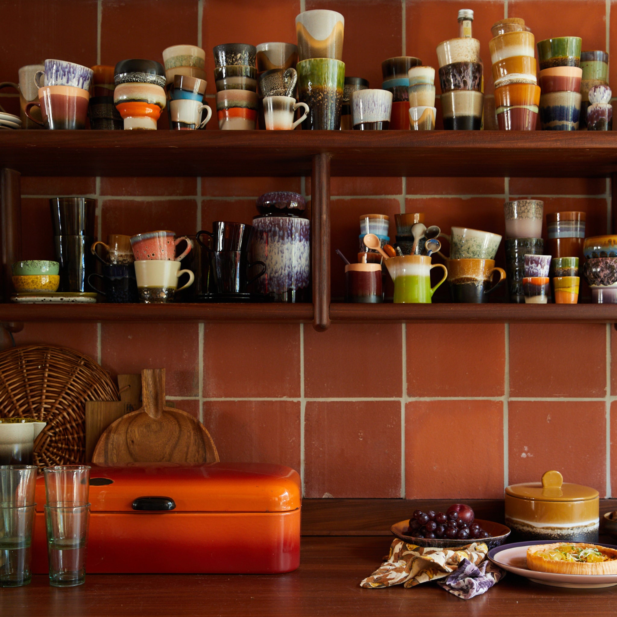 Een gezellige keuken met terracotta betegelde muren, planken met kleurrijke mokken en HK Living 70s Ceramics Eierdop Granite 4 st., houten snijplanken, een fel oranje brooddoos, glaswerk, druiven en een schaal op een houten aanrecht.