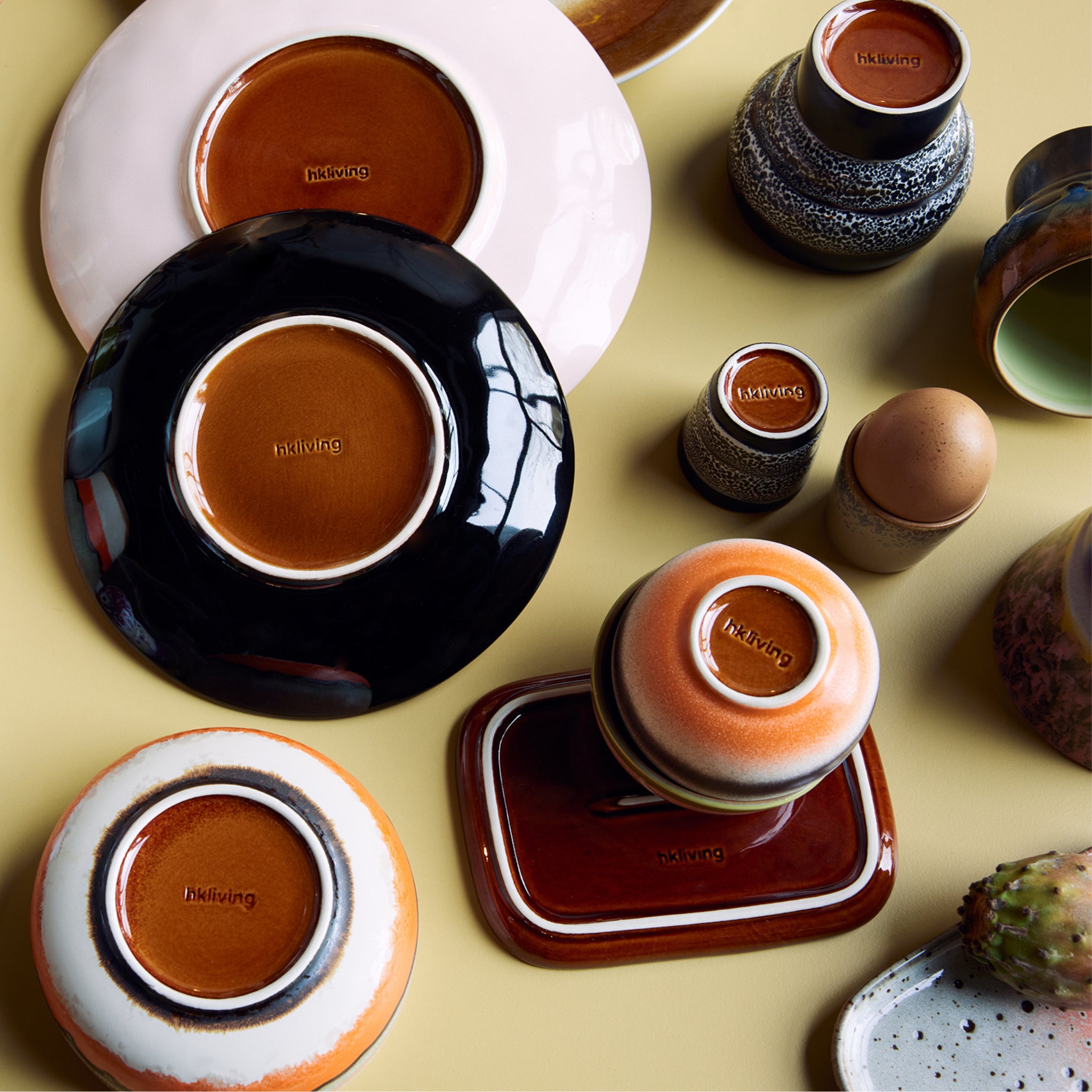 HK Living 70s Ceramics Eierdop Granite 4 st. (eierdopjes) van HK Living worden ondersteboven tentoongesteld met andere borden, kommen en kopjes in verschillende kleuren en texturen op een lichtgeel oppervlak, elk gestempeld "hk living" op de basis.