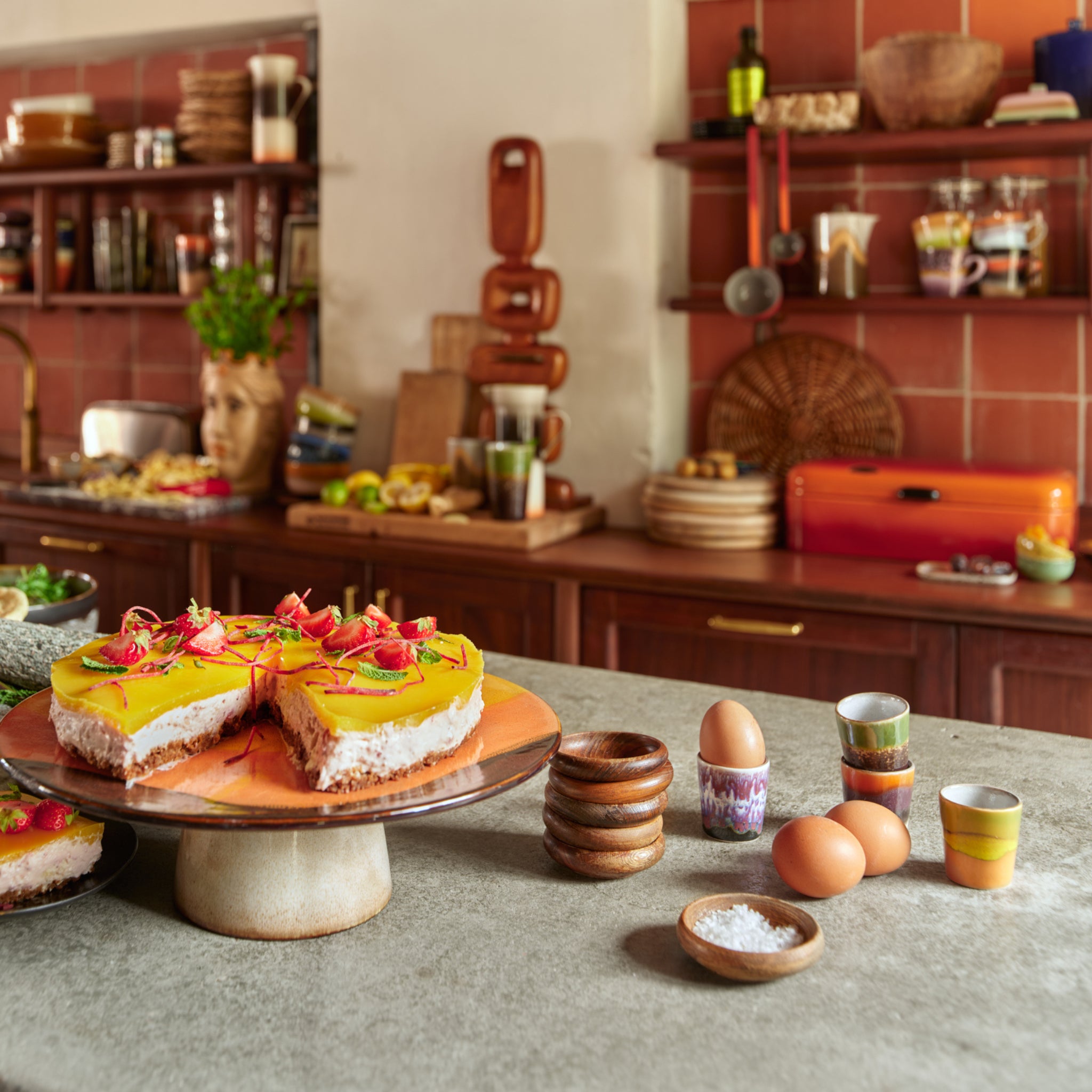 Een keuken met een marmeren aanrecht met daarop een taart met geel glazuur en eetbare bloemen, eieren, zout, houten kommen en een HK Living 70s Ceramics Eierdop Island 4 st. set. Planken achter tonen servies en retro keukengerei.