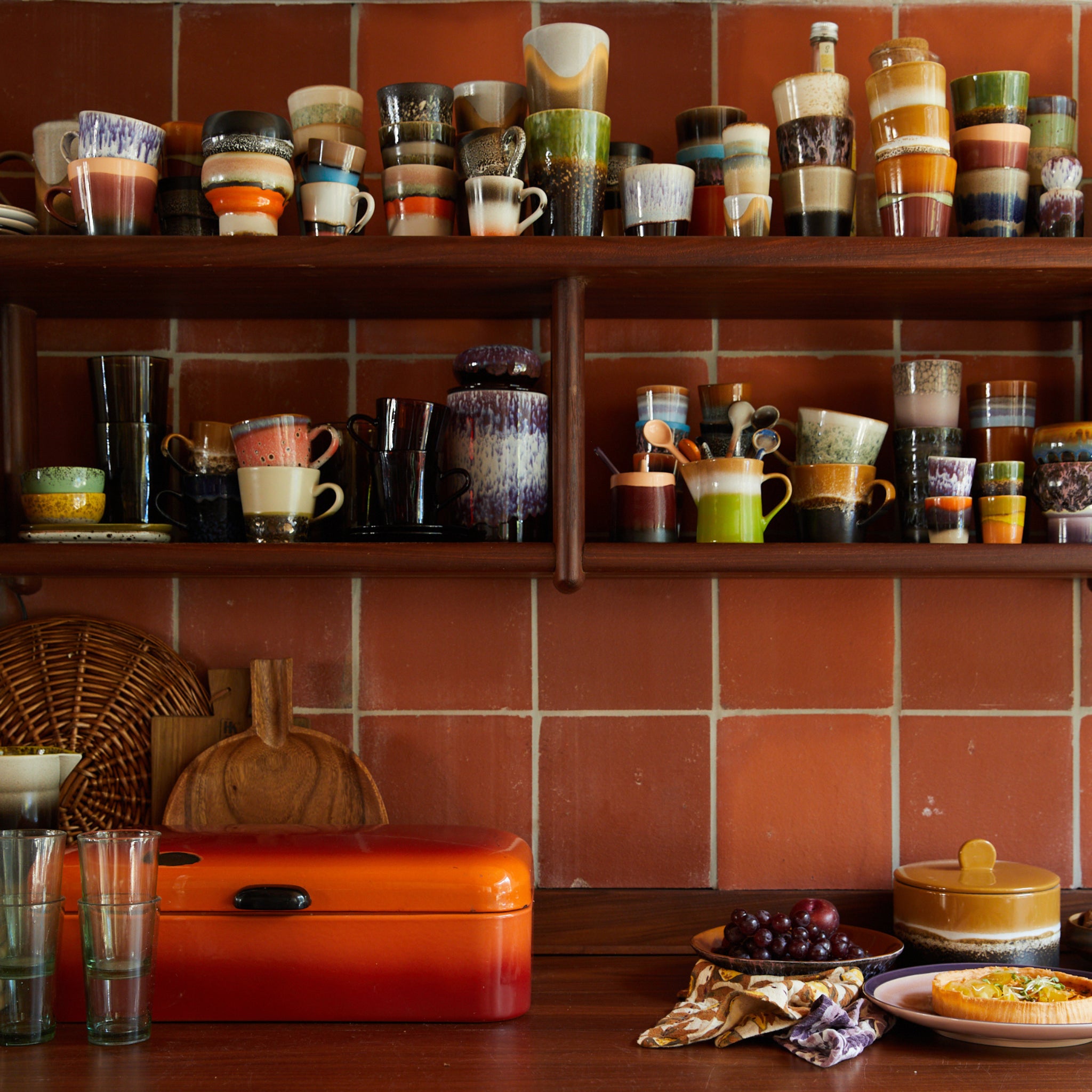 Een keuken met terracotta betegelde muren, houten planken met daarop HK Living 70s Ceramics Koffiekop Algae mokken van HK Living, een rode broodtrommel, glaswerk, snijplanken en een aanrecht met fruit en servies in retrostijl.