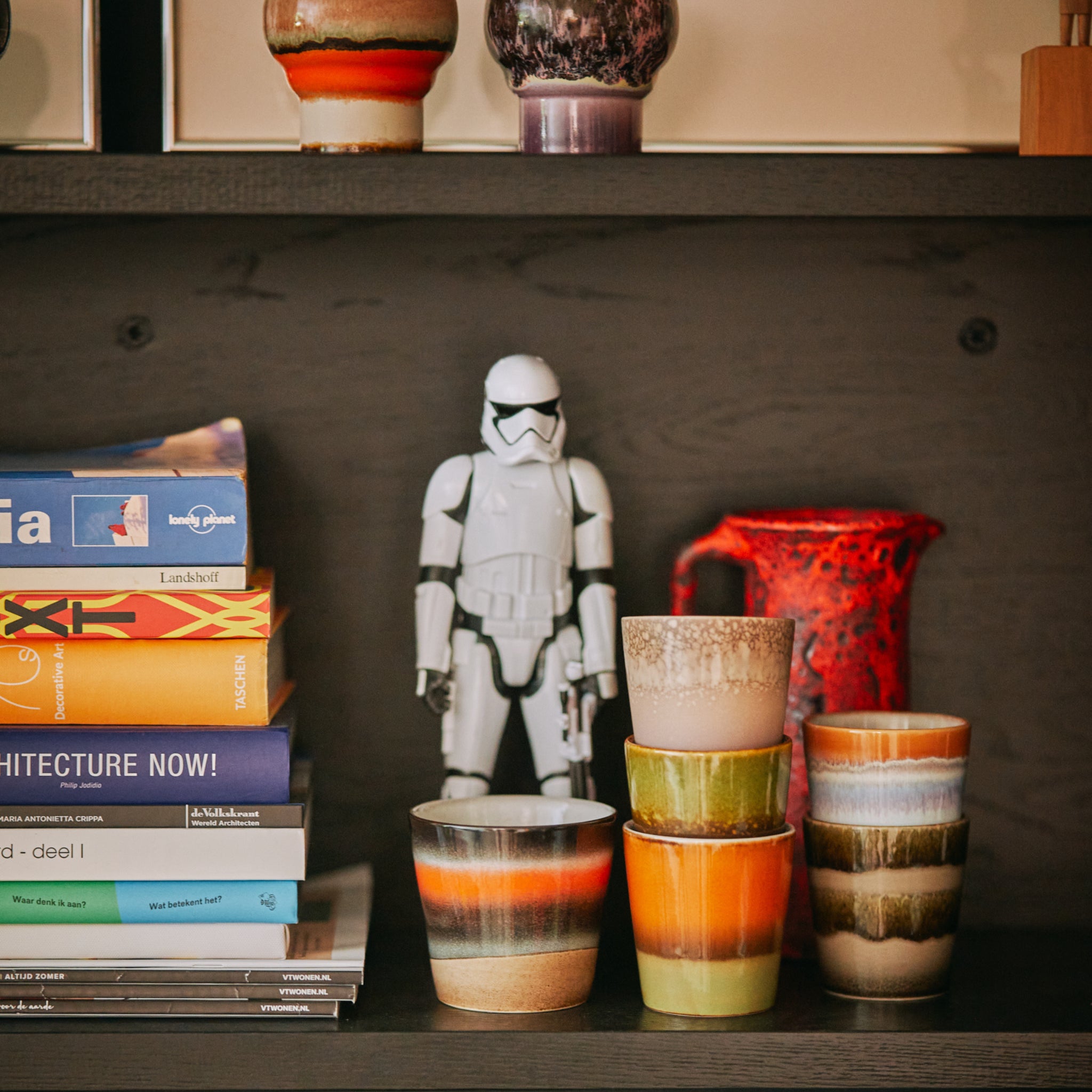 Op een plank staan gestapelde HK Living 70s Ceramics Koffiekop Ash kopjes, een rode kan, een Stormtrooper beeldje en levendige boeken, allemaal in retrostijl tegen een donkerhouten achtergrond.