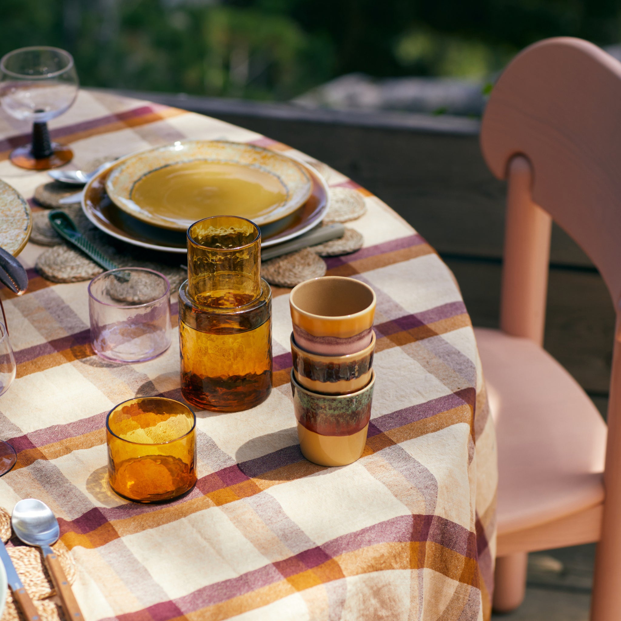 Een tafel gedekt voor een maaltijd met gestapelde amberkleurige glazen, HK Living 70s Ceramics Koffiekop Cliffs mokken en borden op een geruit tafelkleed naast een lichtroze stoel. Het tafereel buiten ademt retrostijl uit met weelderig groen op de achtergrond.