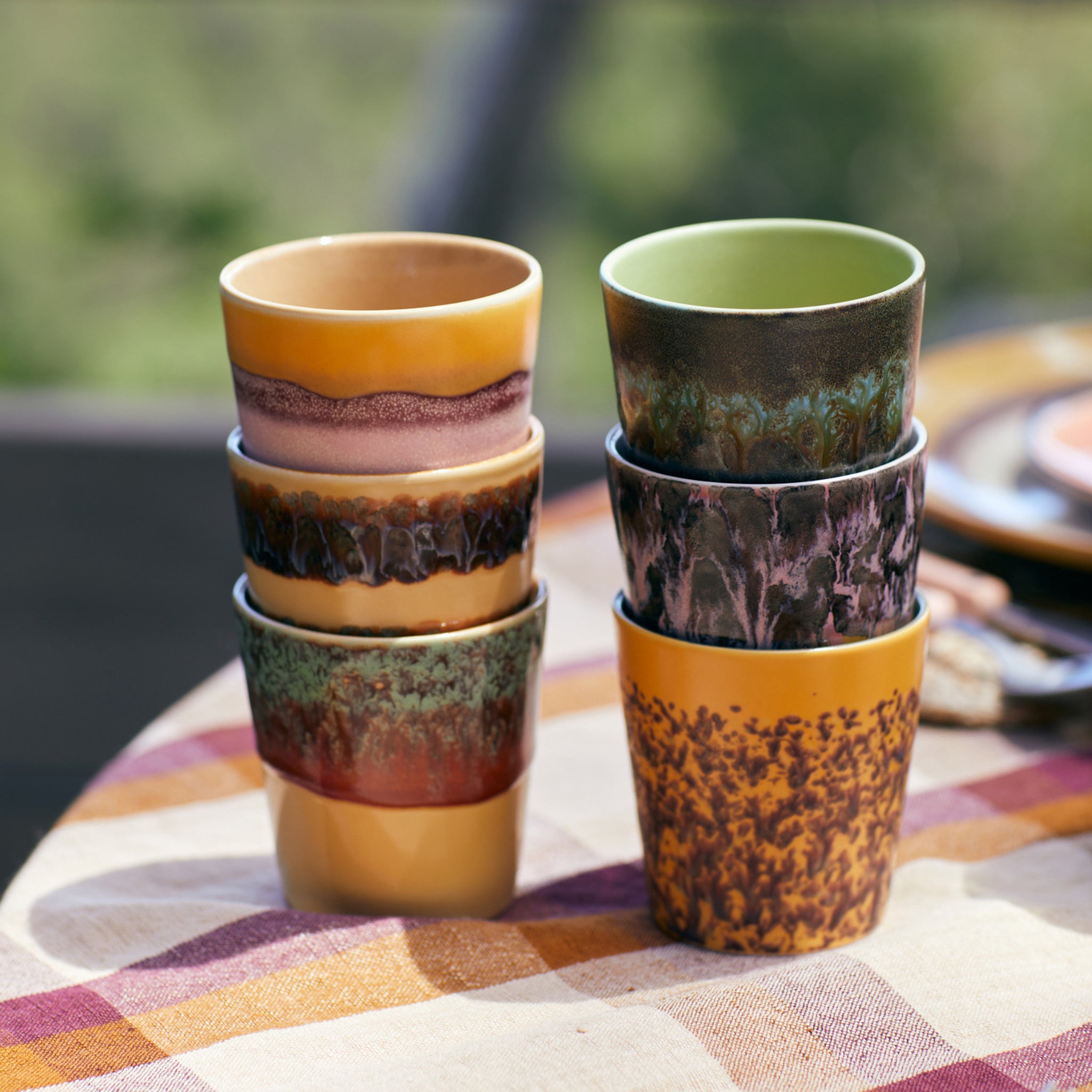 Zes HK Living 70s Ceramics Koffiekop Cliffs kopjes met unieke glazuurpatronen zijn gestapeld in twee stapels op een geruit tafelkleed, waardoor een retro stijl look ontstaat tegen een zacht vervaagde groene achtergrond.