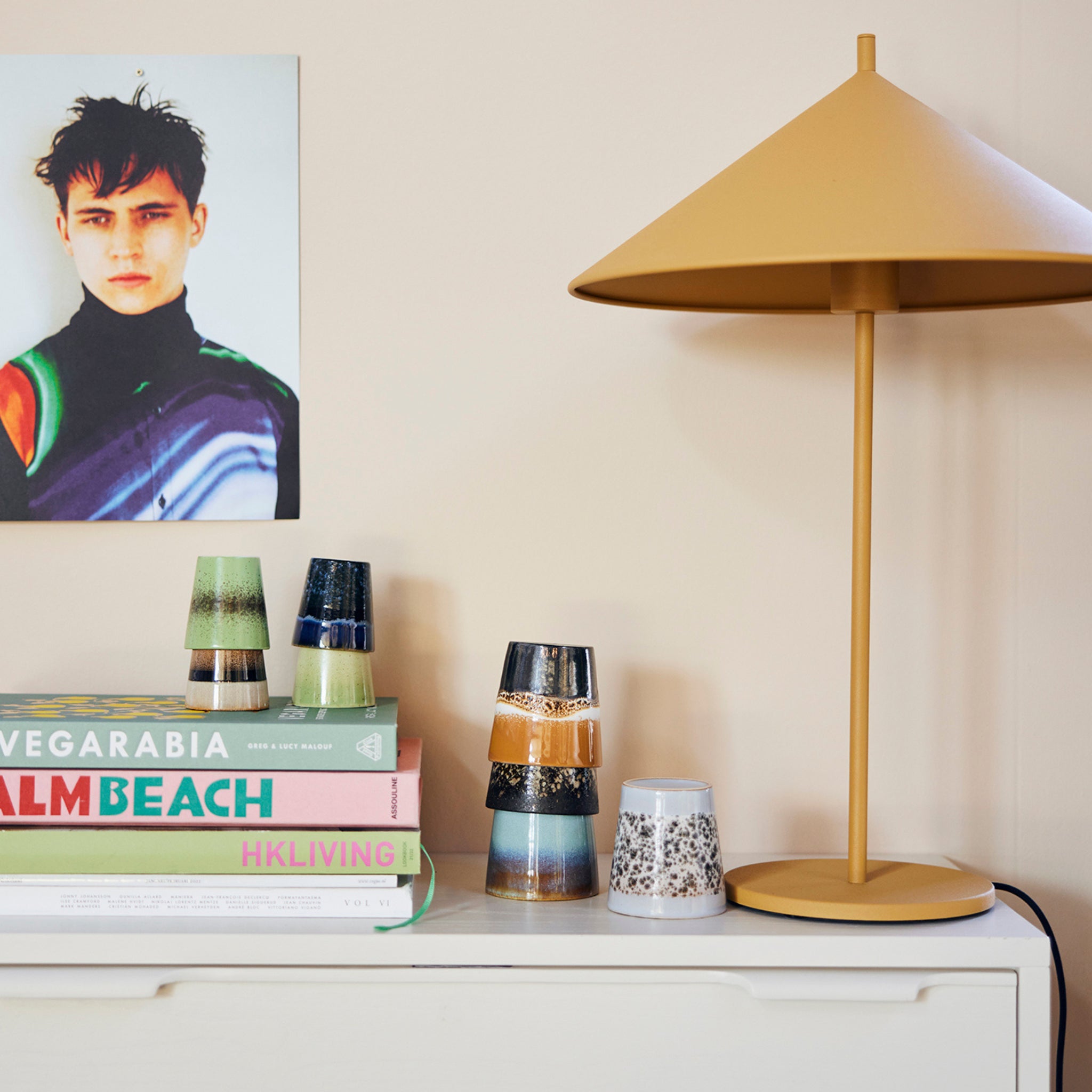 Een gele lamp staat op een wit dressoir naast gestapelde boeken, een HK Living 70s Ceramics Koffiekop Fire mok, kleurrijke kopjes en een muurschildering met een jong persoon met donker haar.