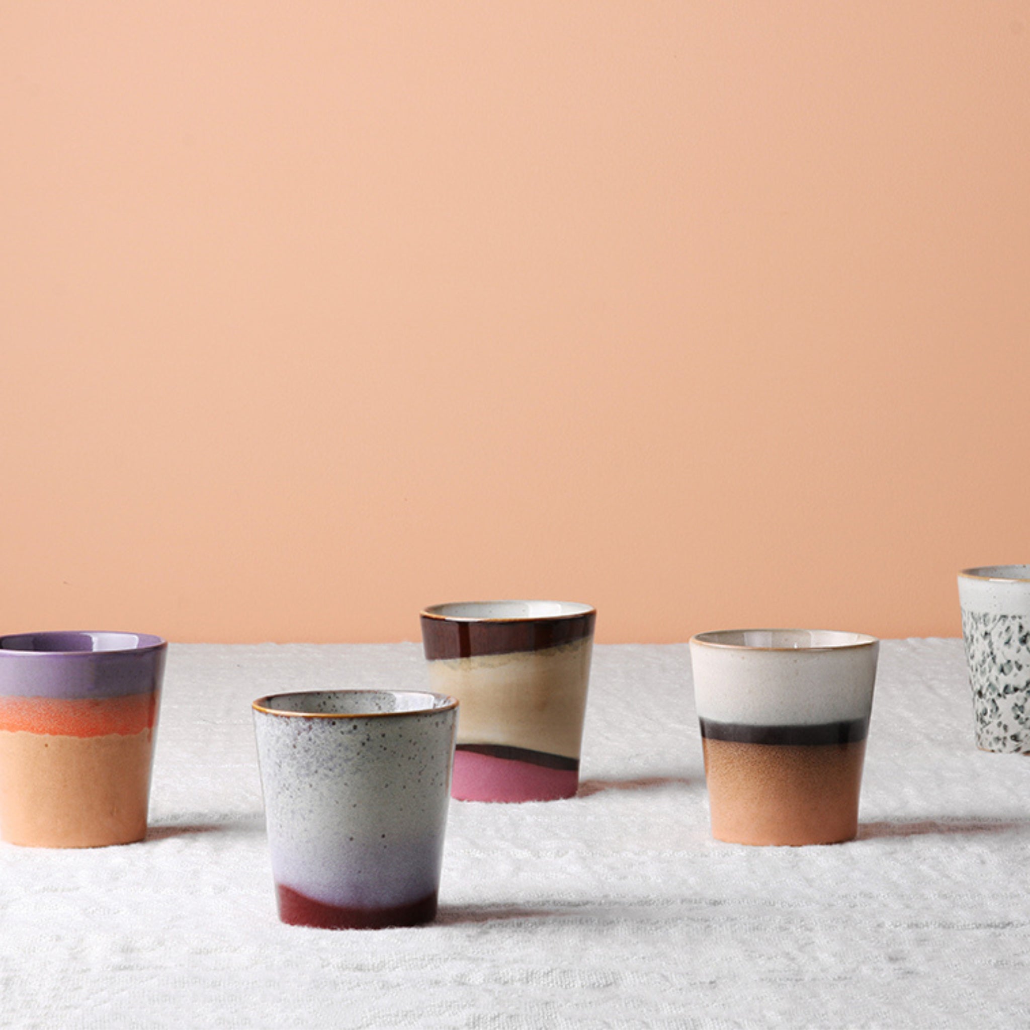 Vijf HK Living 70s Ceramics Koffiekop Frost kopjes, elk met unieke veelkleurige ontwerpen, worden weergegeven op een wit oppervlak met textuur tegen een perzikkleurige achtergrond, die de retrostijl van HK Living keramiek laat zien.