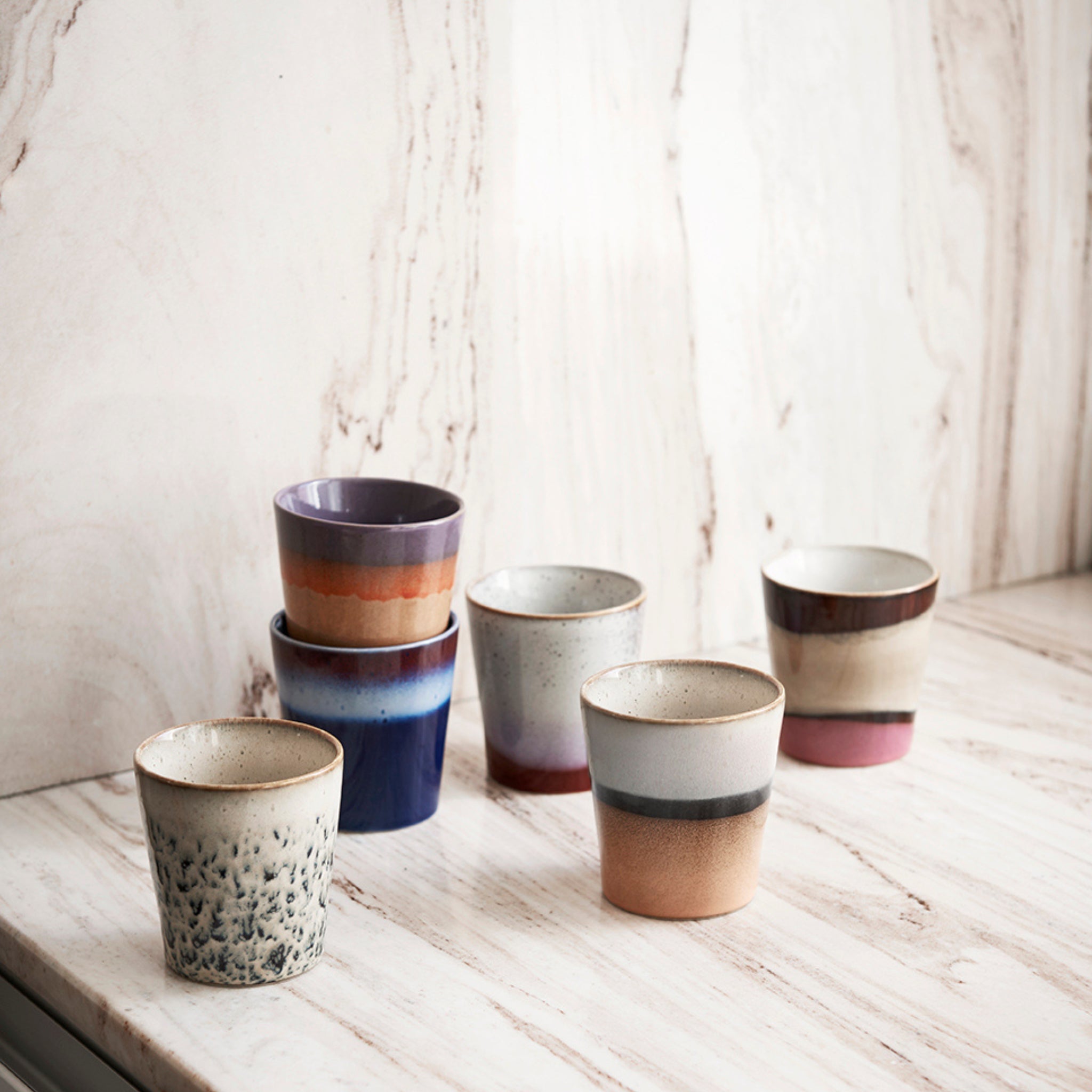Zes HK Living 70s Ceramics Koffiekop Frost-kopjes in retrostijl, elk met unieke kleuren en patronen, zijn gerangschikt op een licht marmeren oppervlak en staan tegen een bijpassende marmeren muur, waardoor hun vintage charme wordt benadrukt.