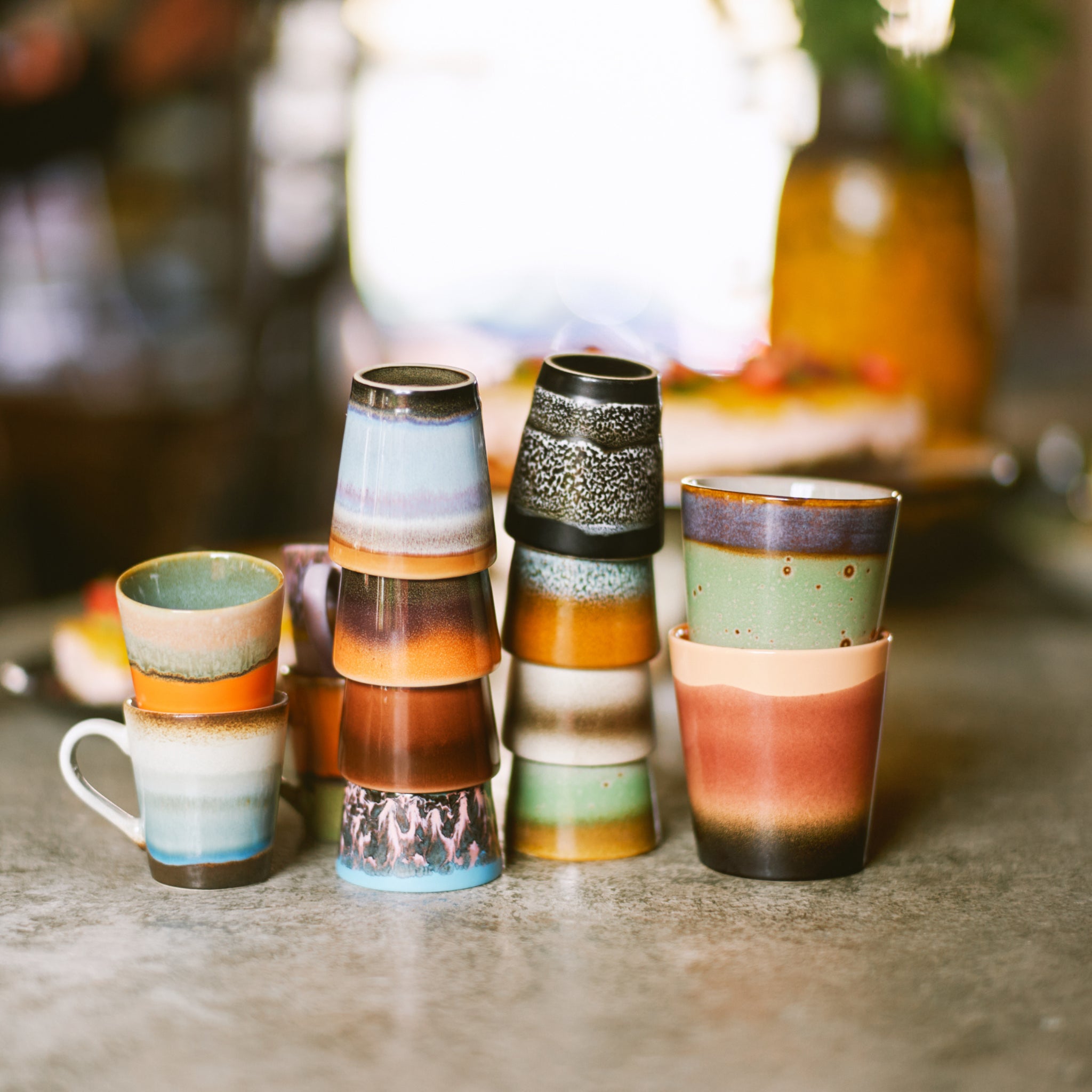 Een stapel kleurrijke HK Living 70s Ceramics Koffiekop Rise mokken met gevarieerde patronen en aardetinten staat op een grijs oppervlak tegen een zacht vage achtergrond.