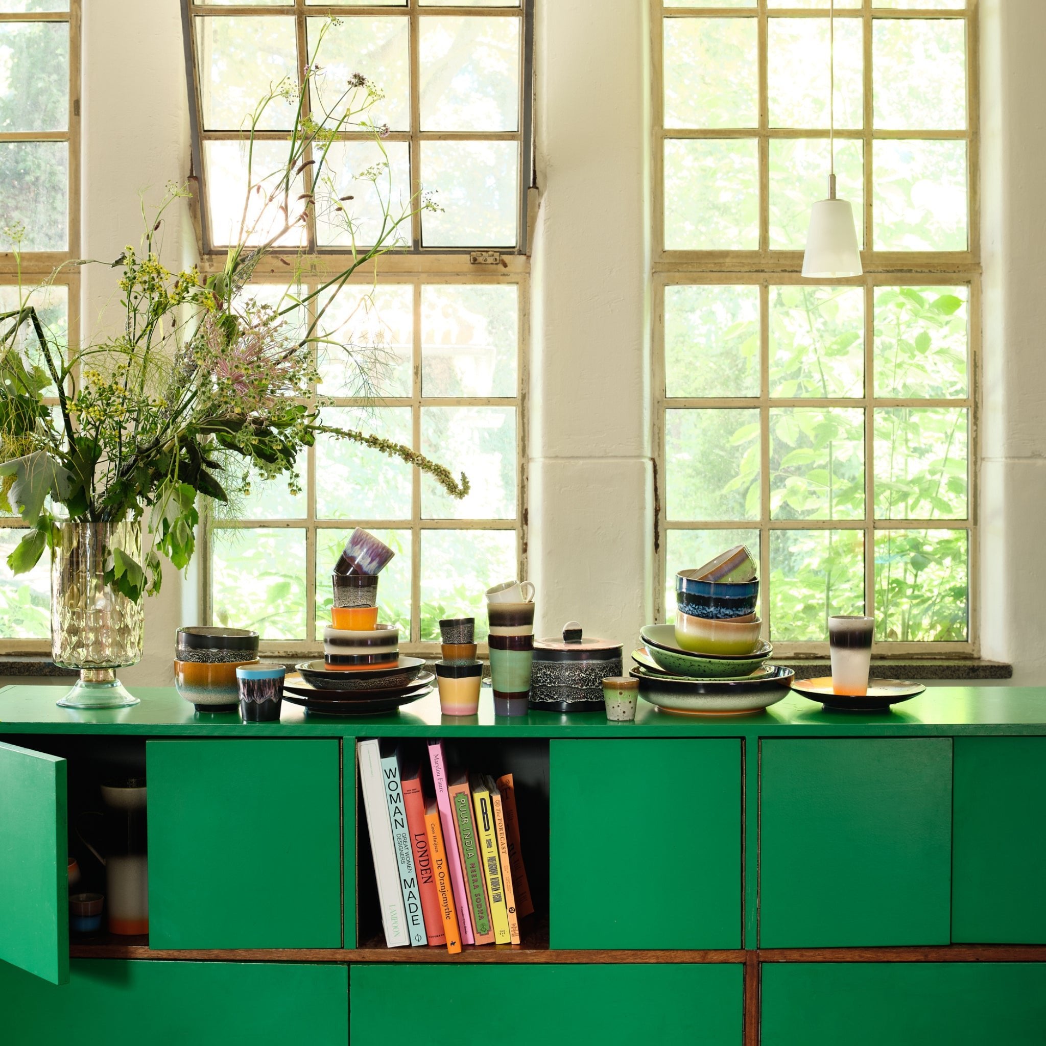 Een groen dressoir met daarop de HK Living 70s Ceramics Koffiekop Yeti en kleurrijke borden staat naast een grote vaas met wilde bloemen. Zonlicht stroomt door de hoge ramen en verlicht de kookboeken in de open kast eronder in retrostijl.
