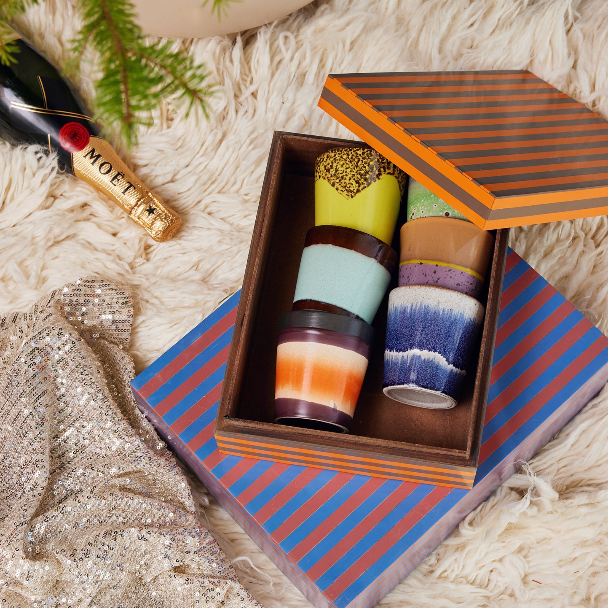 Een open gestreepte doos toont vijf kleurrijke HK Living 70s Ceramics Koffiekop Cosmos mokken op een pluizig tapijt, naast een Moët champagnefles en paillettenstof, met een groene plant in de hoek.