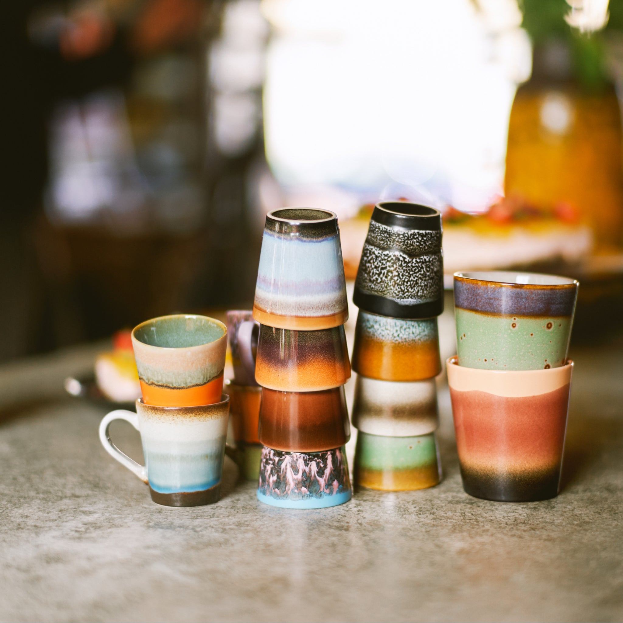 Een set kleurrijke HK Living 70s Ceramics Koffiemok Grounding mokken staat op een tafel, elk met uniek glazuur en aardetinten, met een zachte, onscherpe achtergrond die hun handgemaakte charme benadrukt.