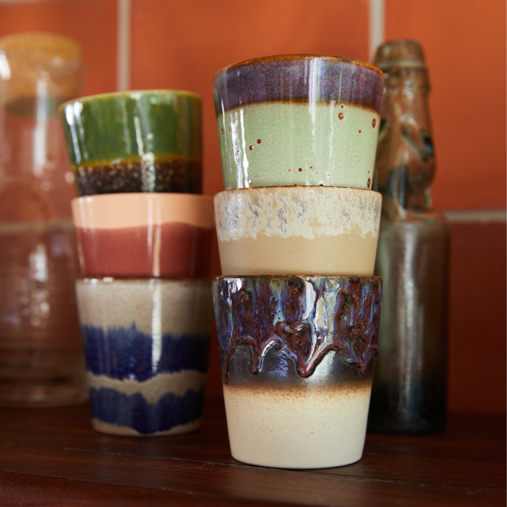 Zes HK Living 70s Ceramics Koffiemok aardingsbekers (set van 6) in verschillende glazuren zijn gestapeld op een houten oppervlak, met glazen flessen en een rode betegelde muur op de achtergrond.