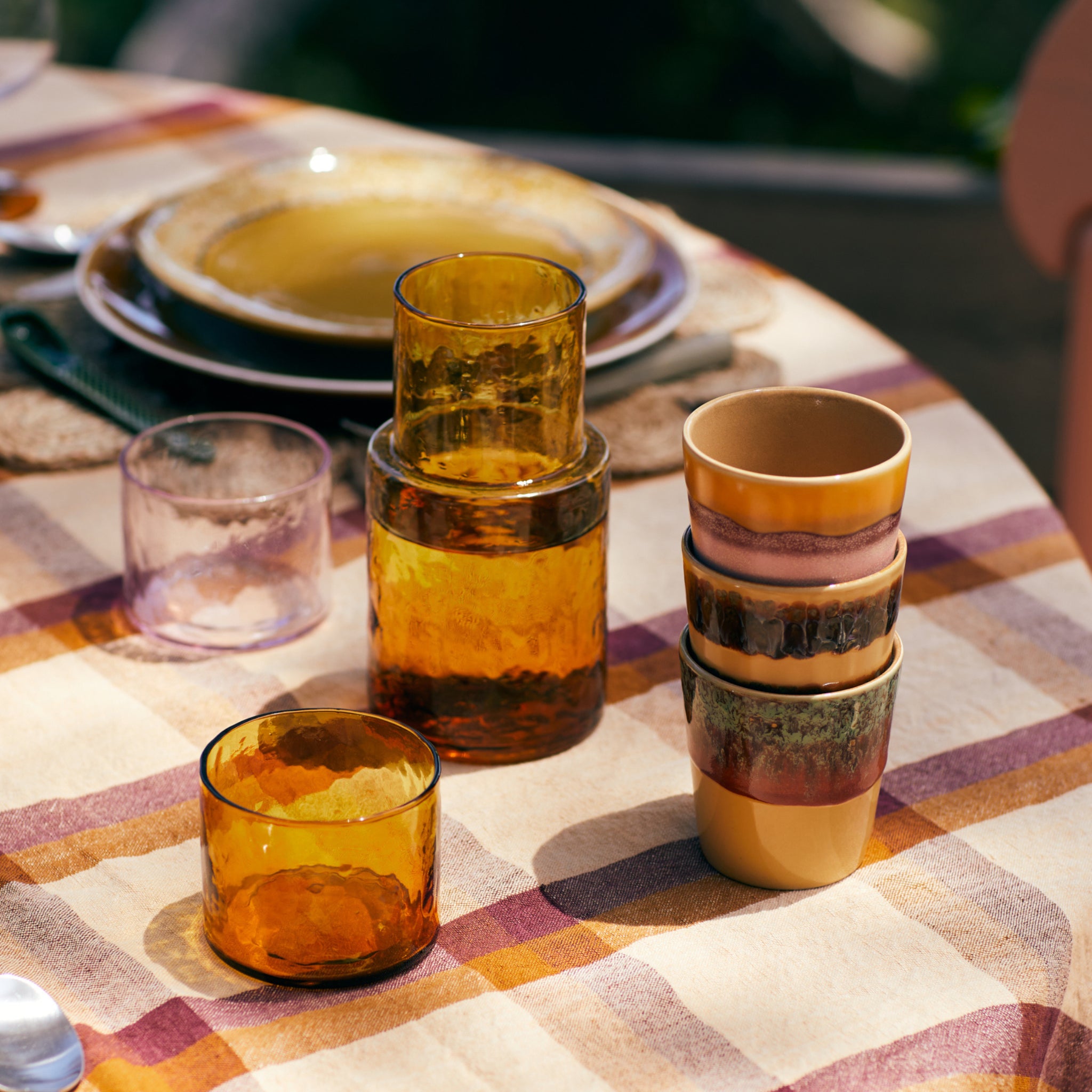 Een close-up van een gedekte tafel met aardse tinten: HK Living 70s Ceramics Koffiemok Valley (set van 6) bovenop andere kopjes, amberkleurig glaswerk en borden op een geruit tafelkleed terwijl het zonlicht zachte schaduwen werpt op het tafereel.