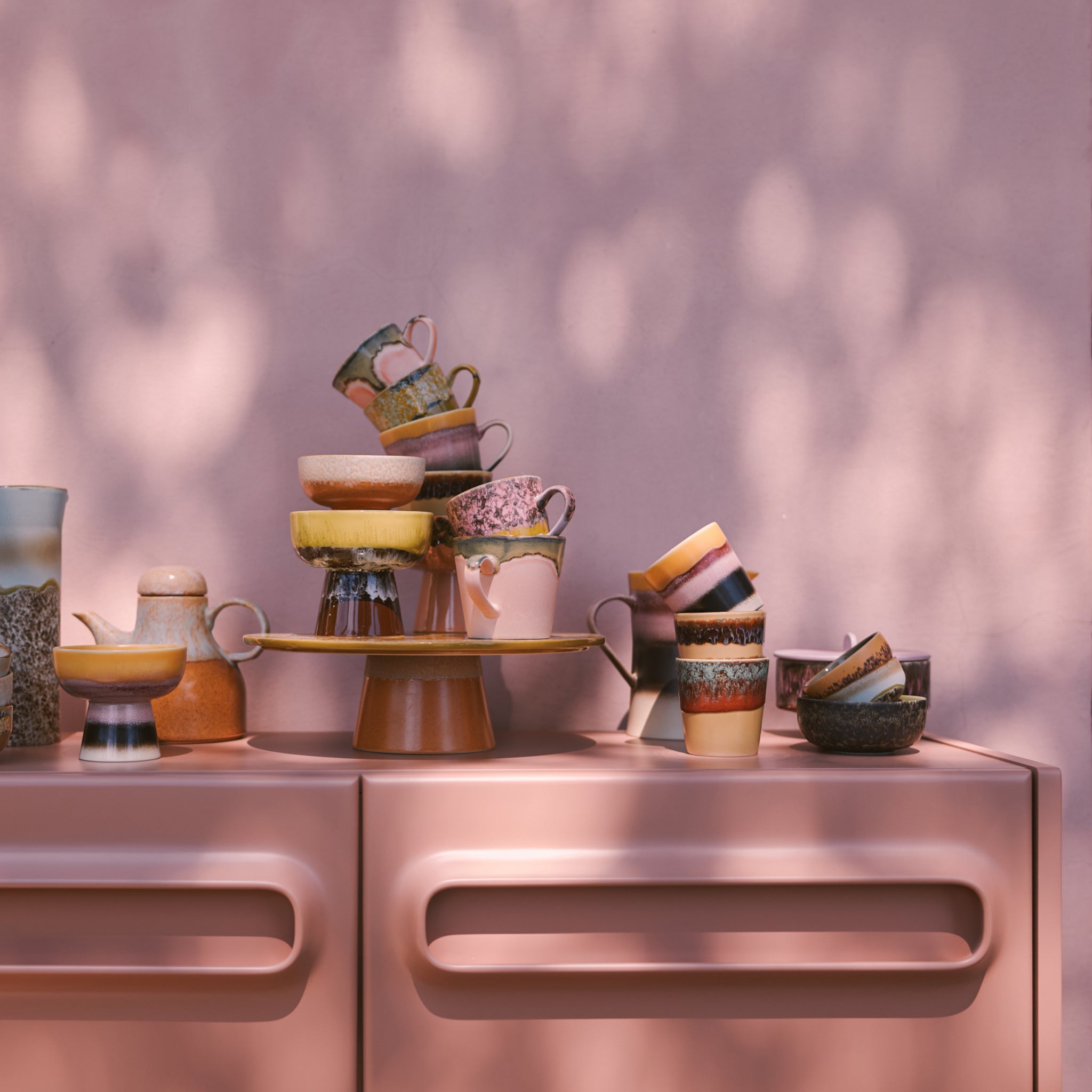Zes HK Living 70s Ceramics Koffiemok Valley mokken staan op een roze kast tegen een roze muur, met gedempt zonlicht en schaduwen van bomen die hun aardetinten accentueren.