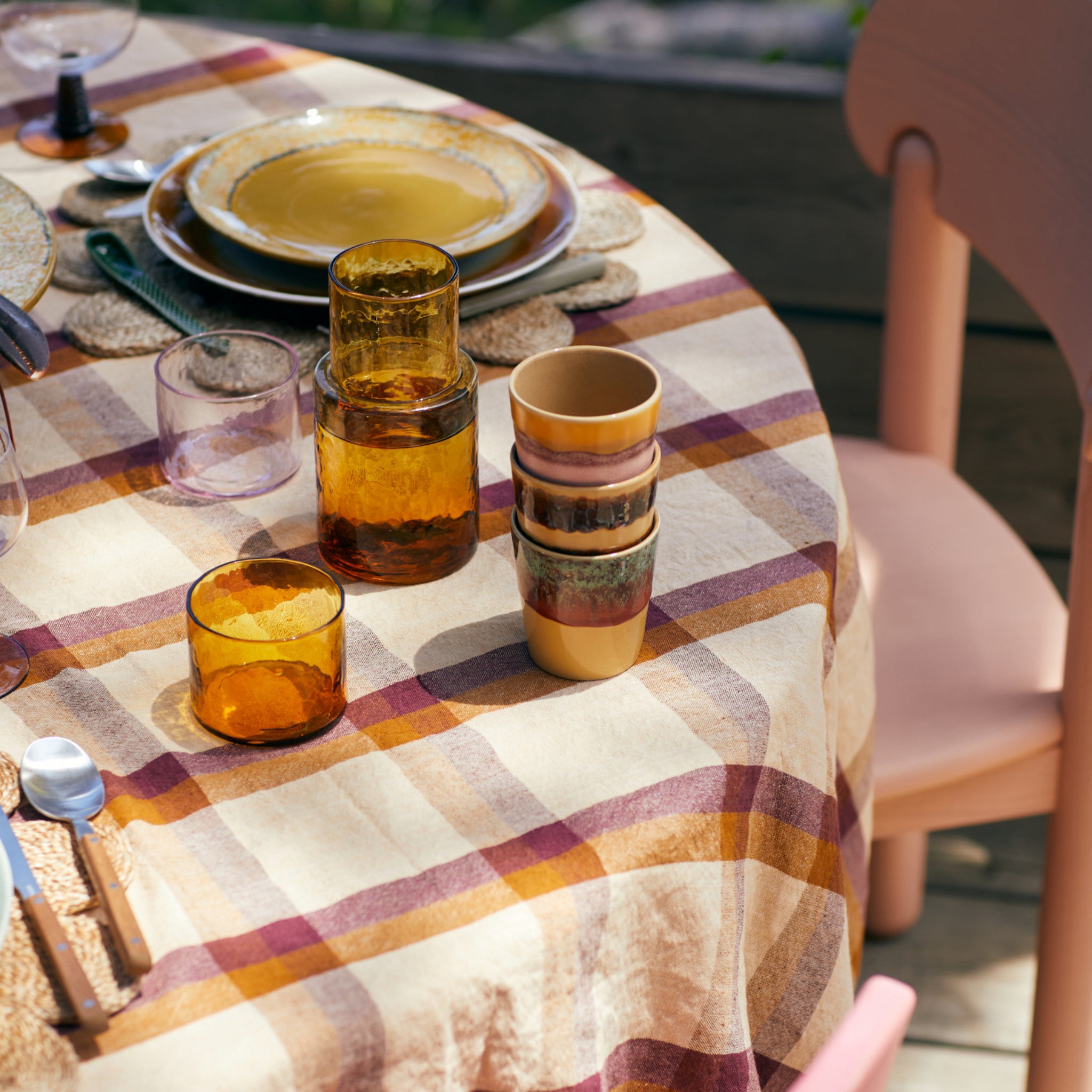 Een ronde tafel buiten in helder daglicht met gele borden, gestapelde bruine en gele glazen en de HK Living 70s Ceramics Koffiekop Cove. Het geruite tafelkleed en de roze stoelen geven het geheel een retro stijl tintje.