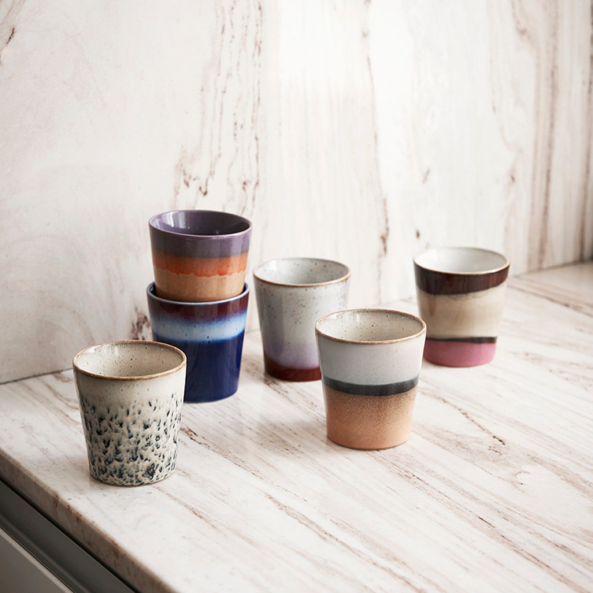 Zes HK Living 70s Ceramics Koffiekop Hail-kopjes in verschillende aarde- en pasteltinten staan op een licht marmeren aanrecht met een marmeren achterwand, wat retro flair geeft aan de omgeving.