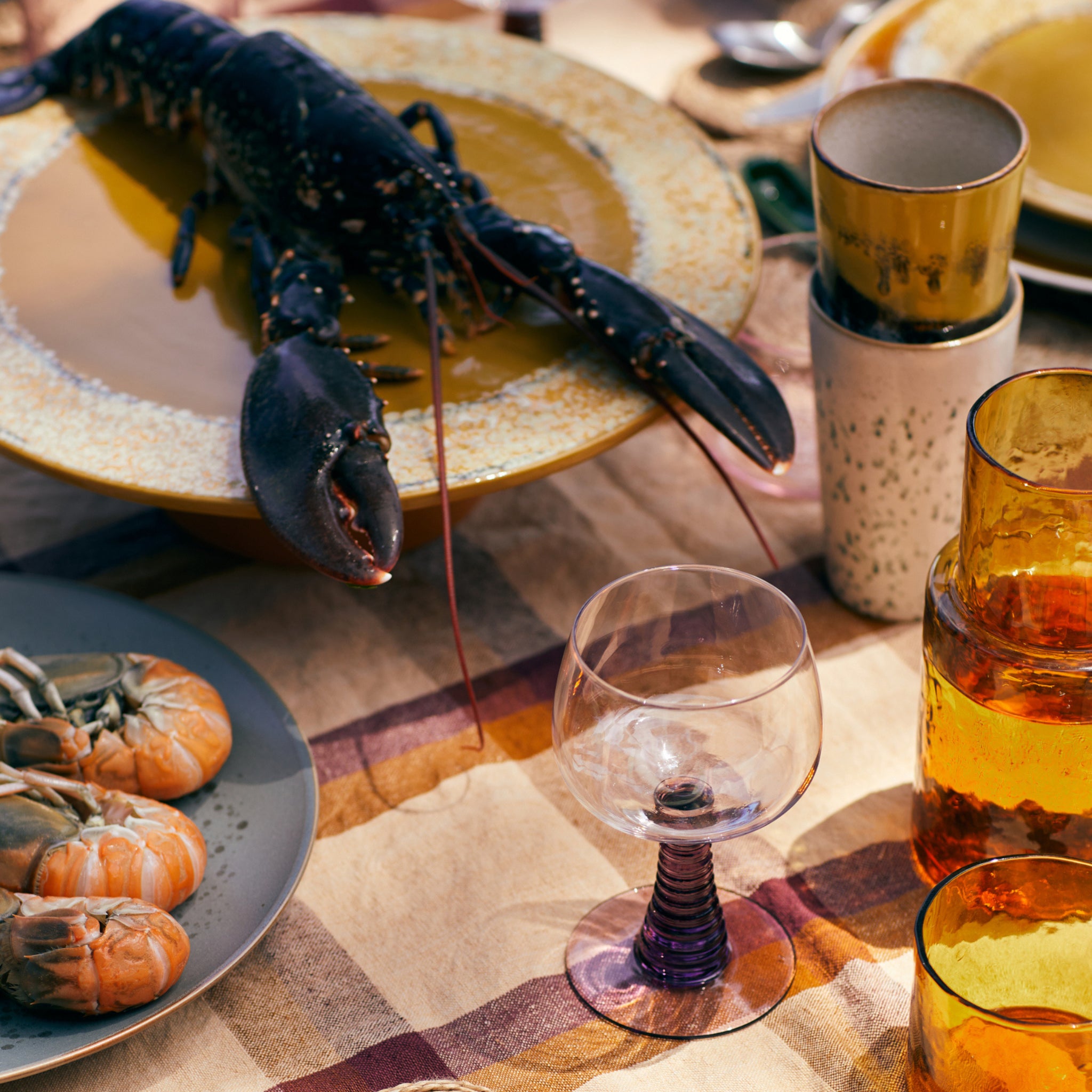 Een gedekte tafel voor een maaltijd bevat een kreeft op een geel bord, garnalen op grijze HK Living 70s Ceramics Latte Mok Creek borden, een leeg wijnglas, amberkleurig glaswerk en gestapelde kopjes op een gestreept tafelkleed.