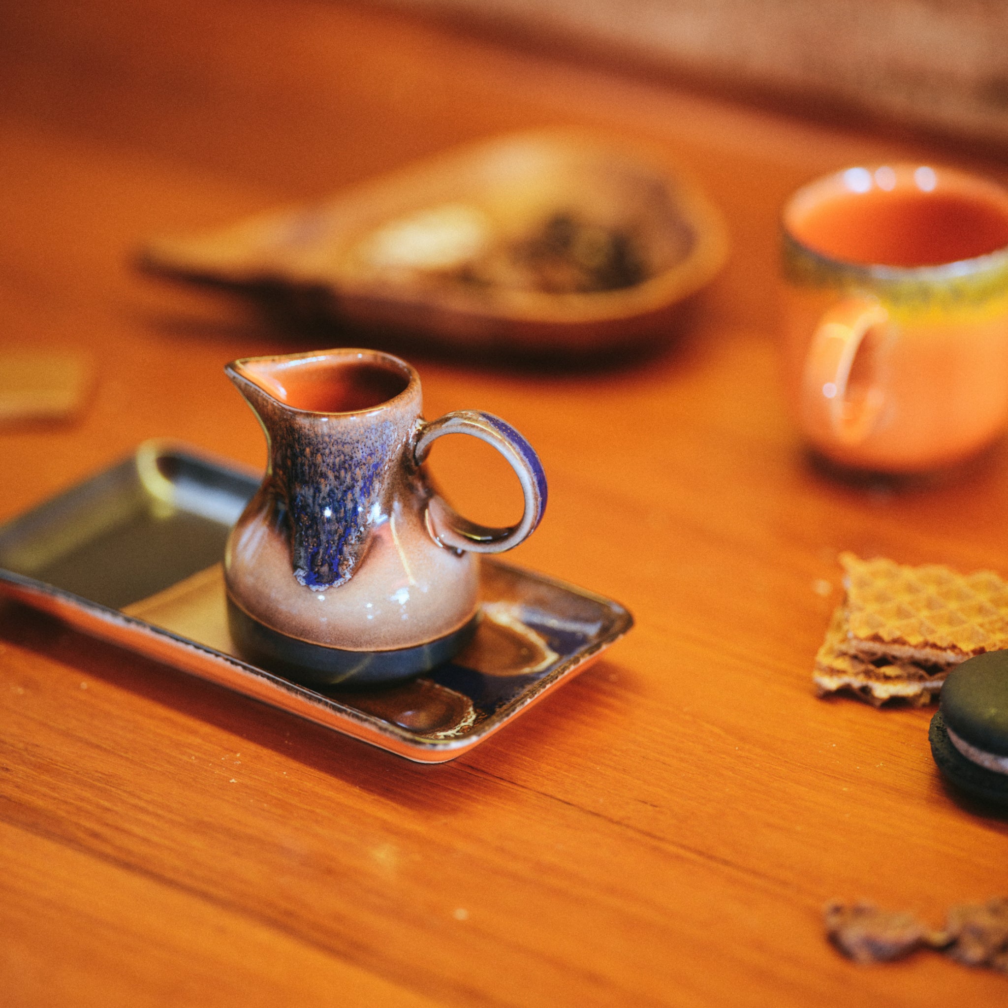 Het HK Living 70s Ceramics Melkkannetje 4PM, een kleine retro creamer met een handvat, staat op een rechthoekig dienblad naast een kopje, houten schaaltje, wafels en donkere macaron op een houten tafel in een warme, uitnodigende scène.