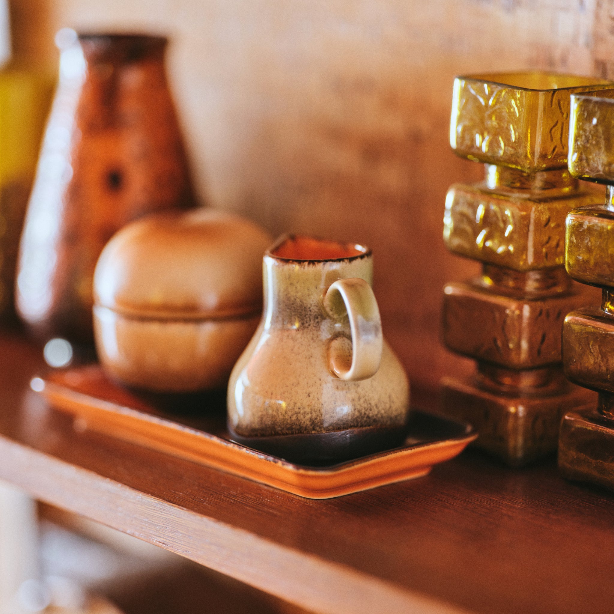 Het HK Living 70s Ceramics Melkkannetje 8AM staat op een oranje dienblad met een kom en gouden blokken, naast ander aardewerk van HK Living, allemaal opgloeiend in warm licht dat doet denken aan klassiek aardewerk uit de jaren 70.