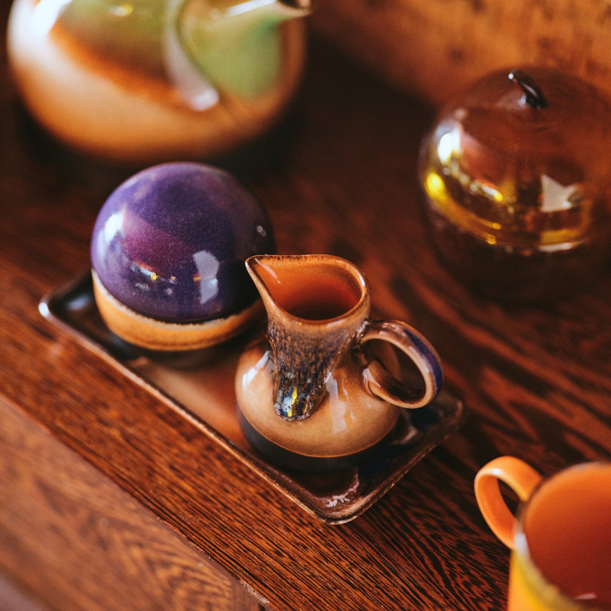 Het HK Living 70s Ceramics Melkkannetje 4PM en een ronde pot met een paars deksel rusten op een rechthoekig dienblad op hout, omringd door ander levendig HK Living keramiek uit de 70s collectie.