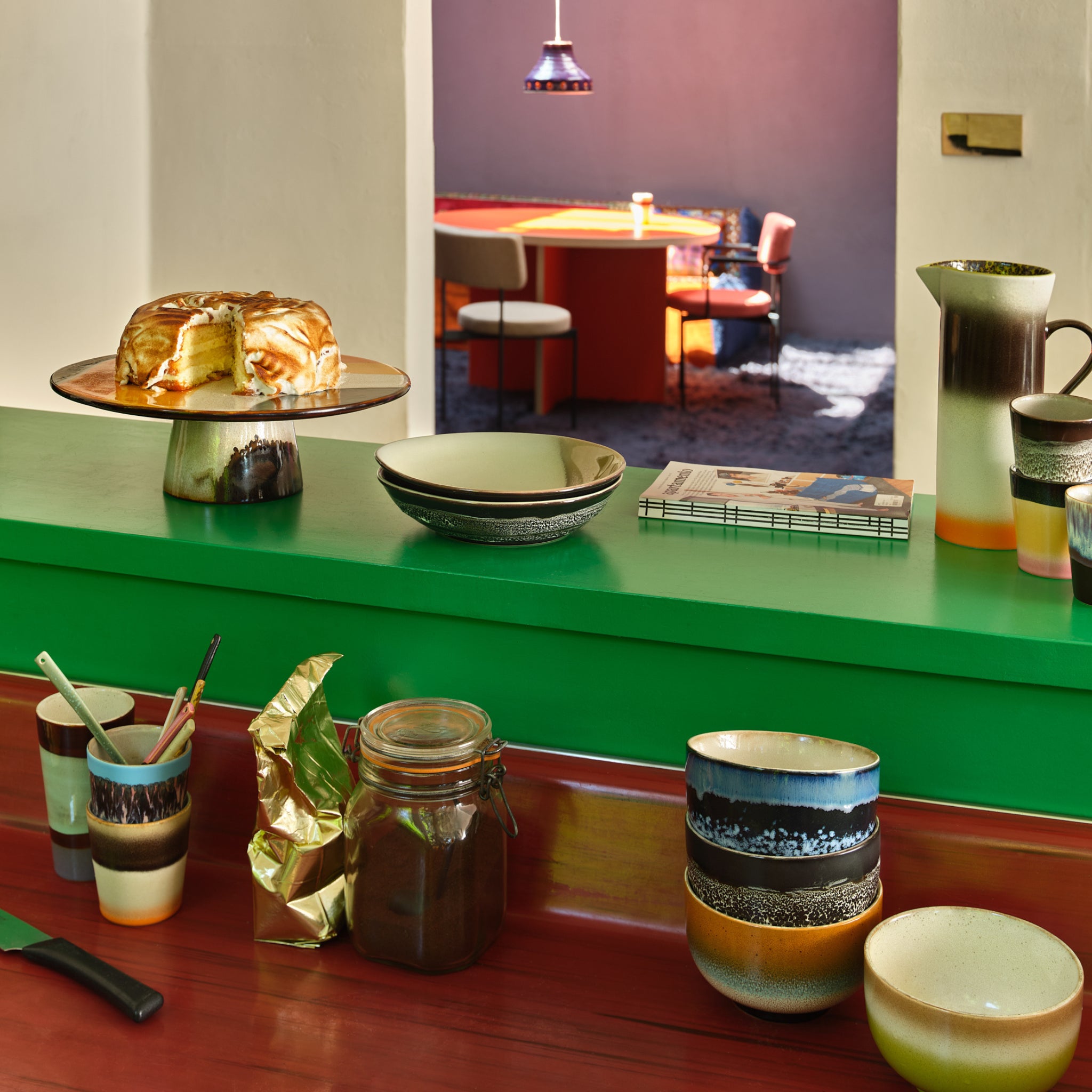 Een levendige keukenscène toont HK Living 70s Ceramics Noodle Kom Groovy D13 kommen, kopjes, een kan, keukengerei, een pot, een ingepakt pakje en cake op een standaard, met een rode tafel en stoelen zichtbaar in de eethoek daarachter.