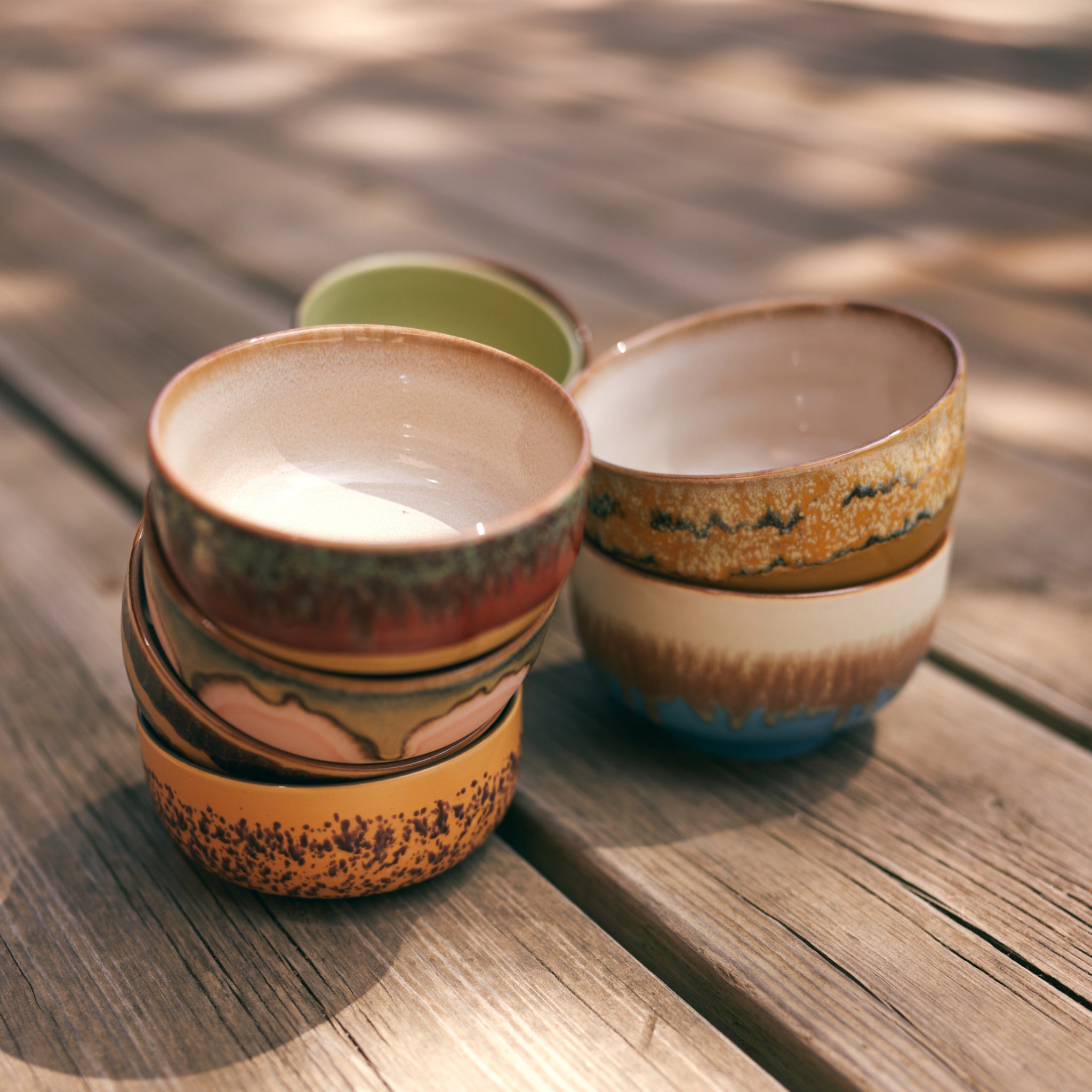 Vier HK Living 70s Ceramics Noodle Kom Seabreeze D13 kommen (set van 4) zijn buiten op een houten oppervlak gestapeld, waarbij het zonlicht hun aardse, geglazuurde patronen en de textuur van zowel de kommen als het hout benadrukt.