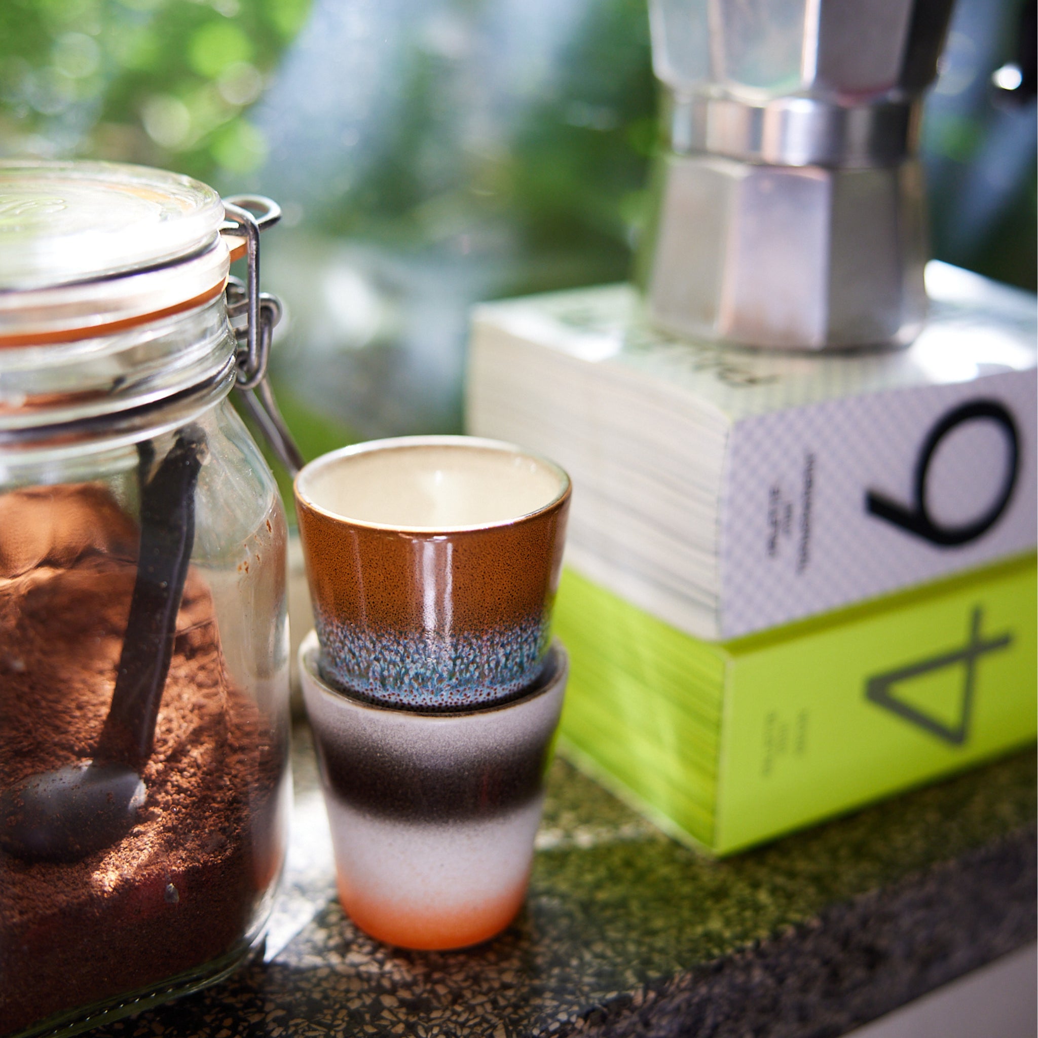 Twee HK Living 70s Ceramics Ristretto Mok Good Vibes kopjes staan opgestapeld naast een glazen pot gemalen koffie met een zwarte lepel, naast een espressomachine op het fornuis en twee boeken op een zonovergoten aanrecht.