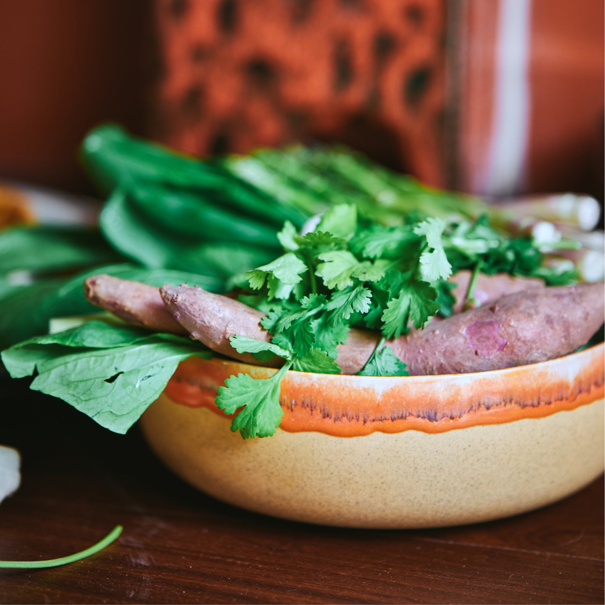 De HK Living 70s Ceramics Slakom Shore D25, met handgemaakte details, staat op een houten oppervlak met bladgroenten, zoete aardappels en koriander erin. Deze unieke keramische kom van HK Living steekt af tegen een vage achtergrond.