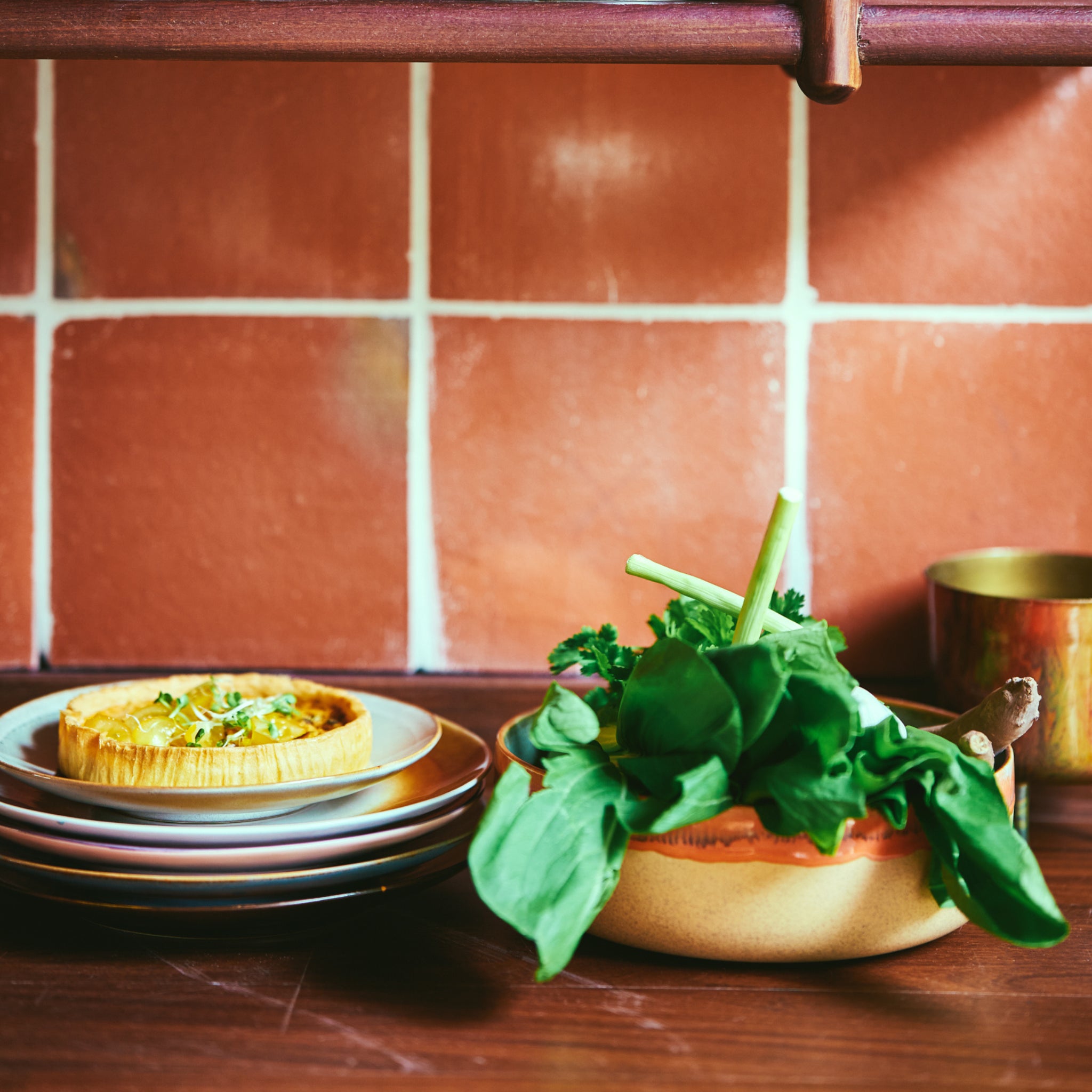 De HK Living 70s Ceramics Slakom Shore D25, gevuld met vers groen, staat op een houten toonbank naast borden en een quiche, waardoor een stijlvol tafereel ontstaat tegen een roodbruine betegelde muur.