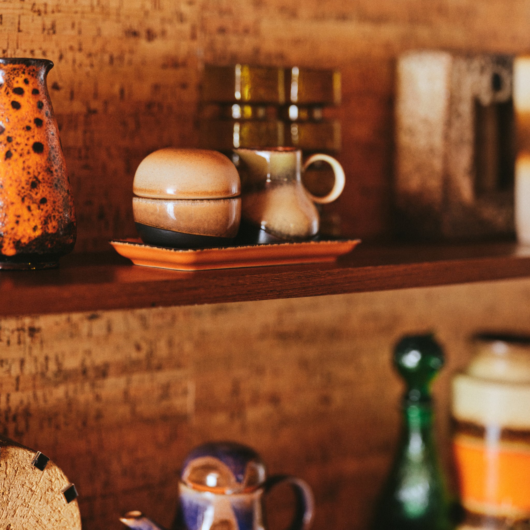 Op een houten plank staan HK Living 70s Ceramics Suikerpot 8AM, vintage mokken, kommen en kopjes in aardetinten. Groene glazen flessen en een bruine wand met textuur roepen een warme, retro sfeer op geïnspireerd door HK Living klassiekers.