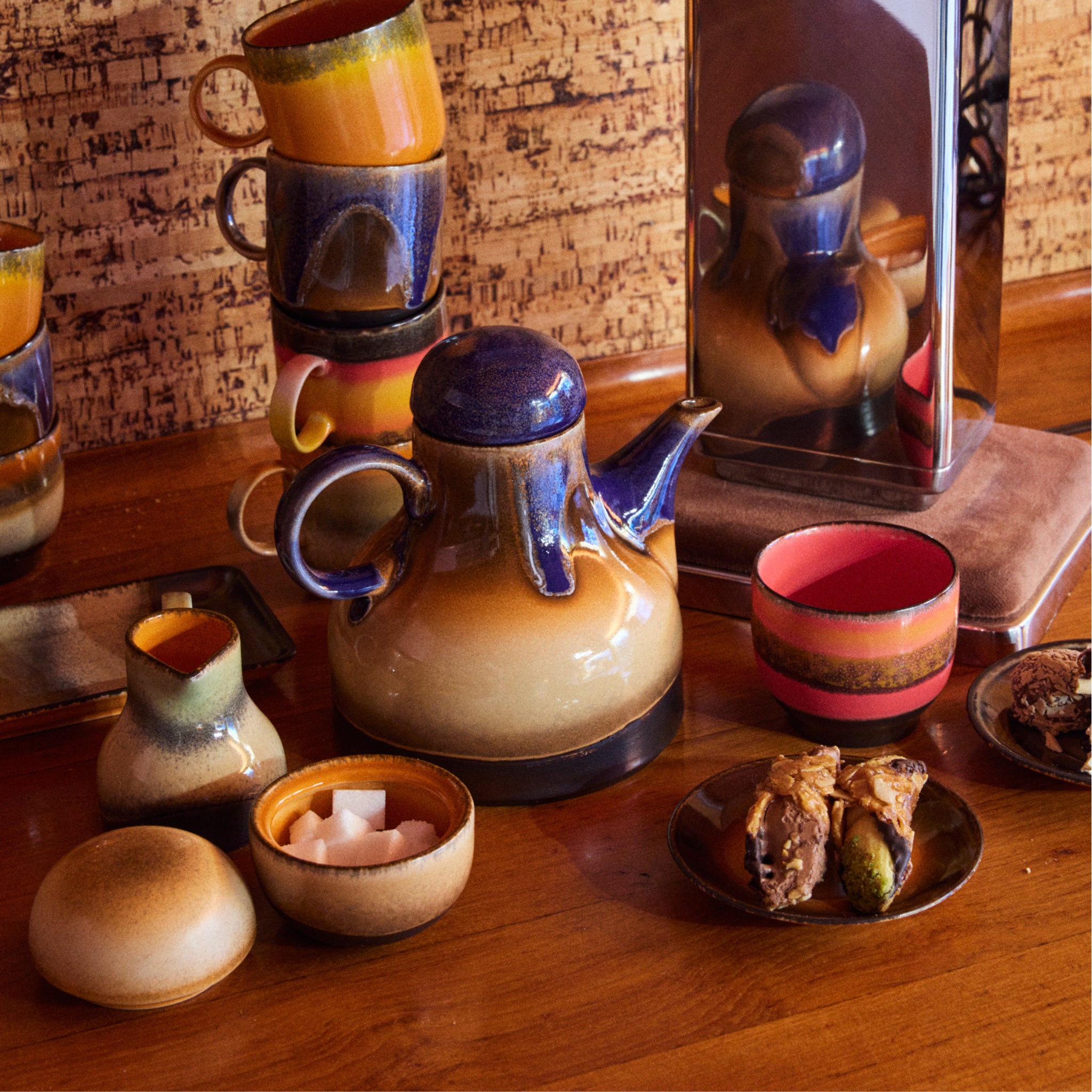De HK Living 70s Ceramics Suikerpot 8AM staat op een houten tafel met bijpassende kopjes, een blauwe en bruine theepot, een kleine kan, baklava op een bord, een roze gestreepte kom en een spiegel op de achtergrond.