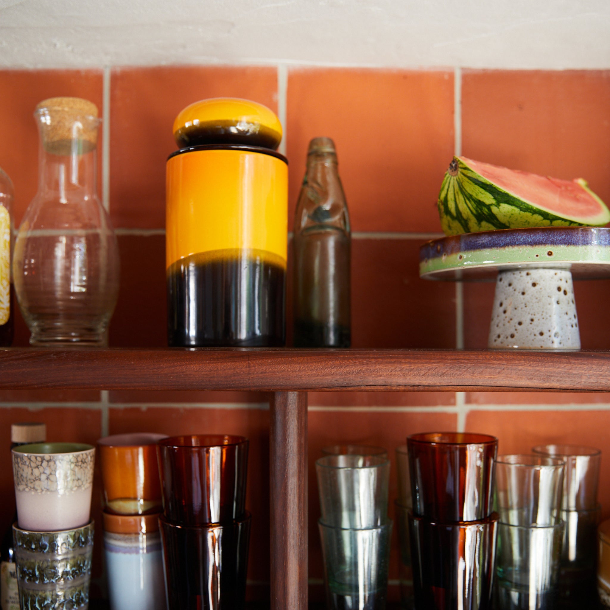 Op een houten plank tegen oranje tegels staan een HK Living 70s Ceramics Voorraadpot Sunshine, een glazen fles en een schijf watermeloen op een keramische standaard. Hieronder staan gestapelde kleurrijke drinkglazen in verschillende ontwerpen.