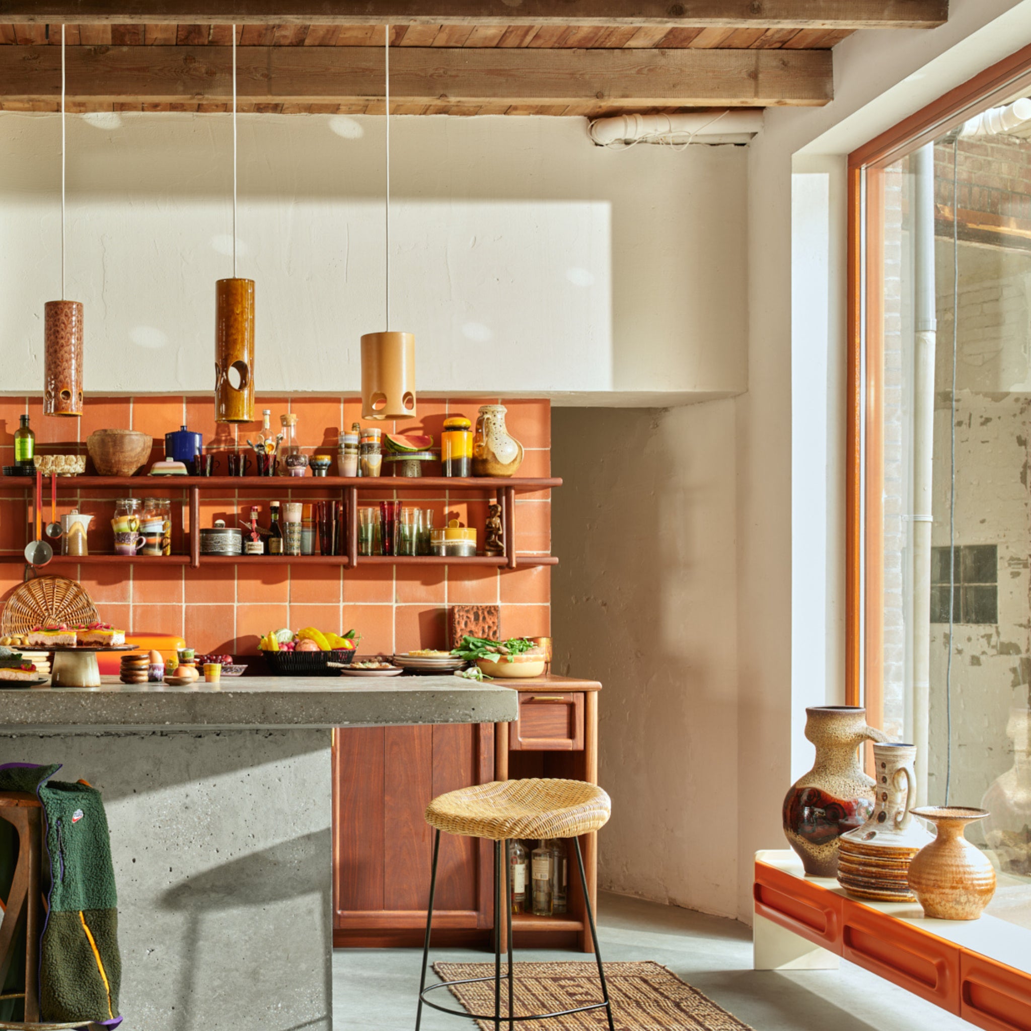 Een gezellige keuken heeft terracotta tegels, houten planken met potten en schalen, een betonnen eiland met eten, een gevlochten kruk en zonlicht dat door een groot raam naast de HK Living 70s Ceramics Voorraadpot Sunshine schijnt.